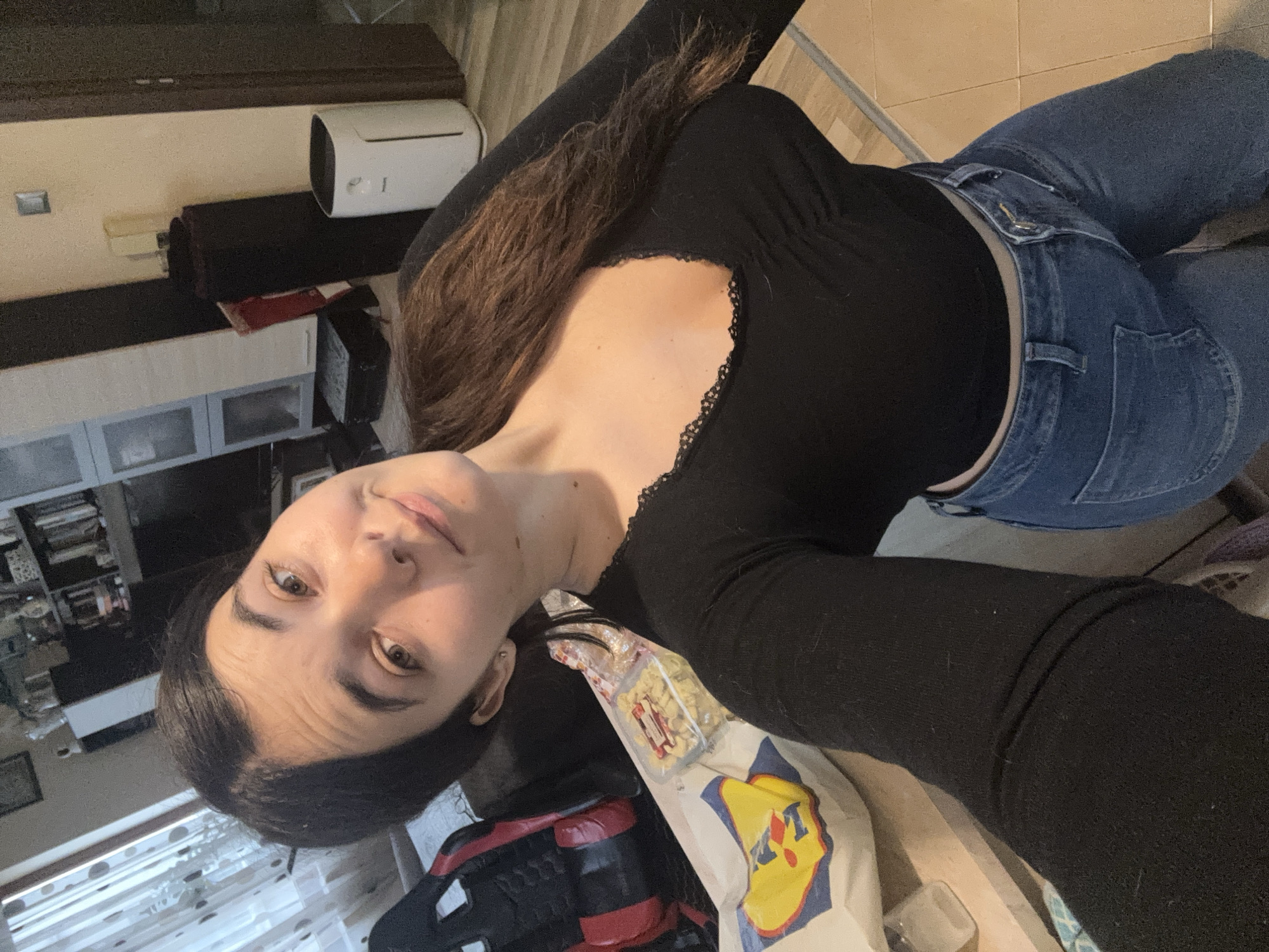 A young woman is captured in a selfie, holding the camera from a high, tilted angle, looking directly into the lens with a neutral expression. She has long, dark brown hair and fair skin. She is wearing a form-fitting black long-sleeve top with a V-neckline adorned with black lace trim, and light blue denim jeans.
The setting appears to be indoors, likely a residential space such as a living room or bedroom. The floor is covered with light-colored tiles. In the background, various household items are visible, including a dark wooden cabinet or shelf unit, white cabinets with glass inserts, and a white, cylindrical appliance that resembles an air purifier or humidifier sitting on a dark surface. To the woman's right, a white shopping bag featuring the "Lidl" logo in yellow and blue is prominently displayed, with a transparent packet of snacks visible inside. Part of a red and black item, possibly a bag or piece of equipment, is also in the foreground.
The lighting suggests it is daytime, with natural light contributing to the illumination of the scene. The visible text is the "Lidl" logo on the shopping bag. The image context is Plovdiv, Bulgaria.