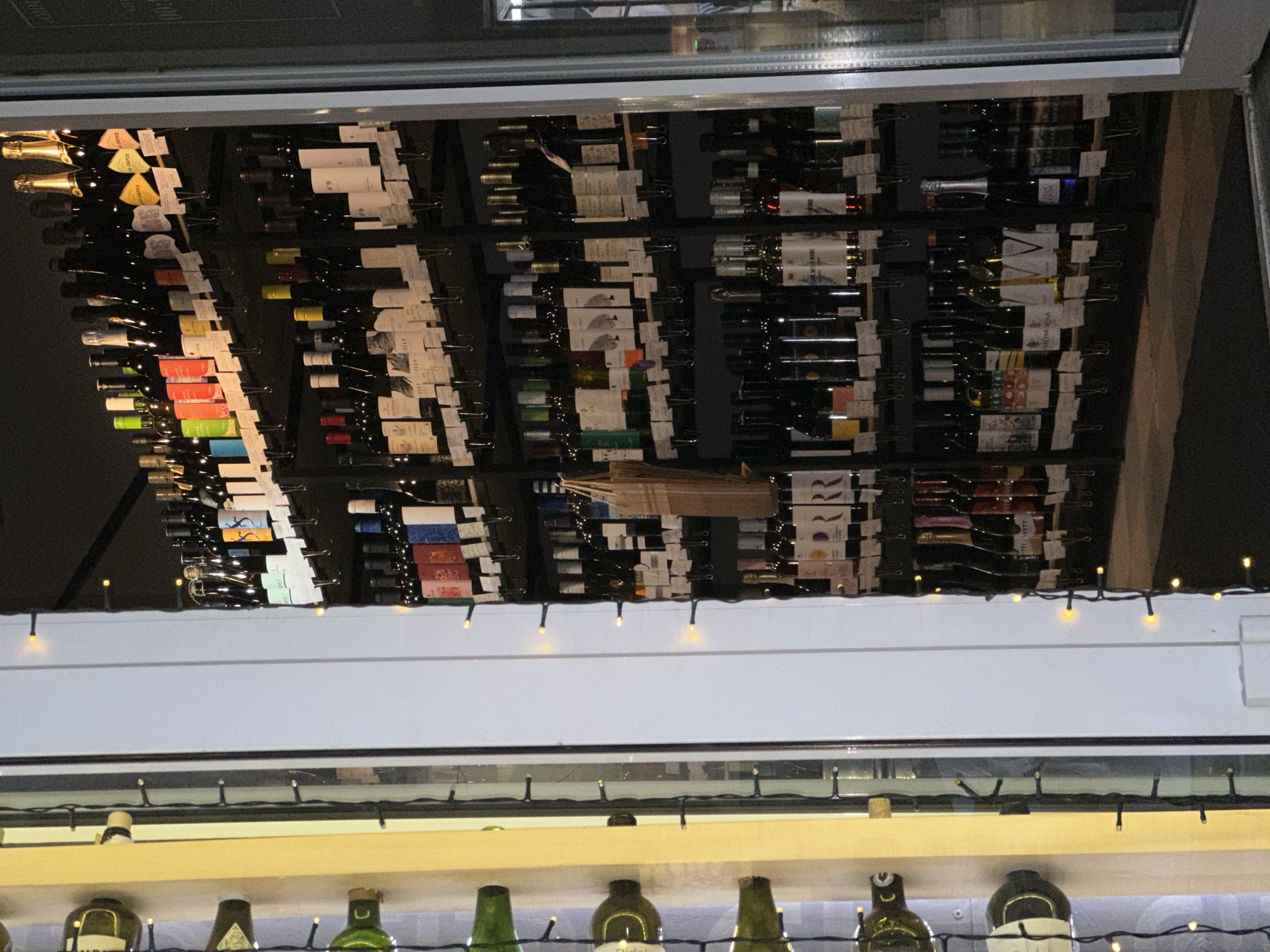 The image captures a wine rack inside a building, possibly a restaurant or a bar in Rostov-on-Don, Russia. The rack is densely populated with wine bottles, each labeled with a tag. Above the rack, a series of lights illuminates the scene. Below the rack, an additional shelf displays bottles with a variety of shapes and colors.