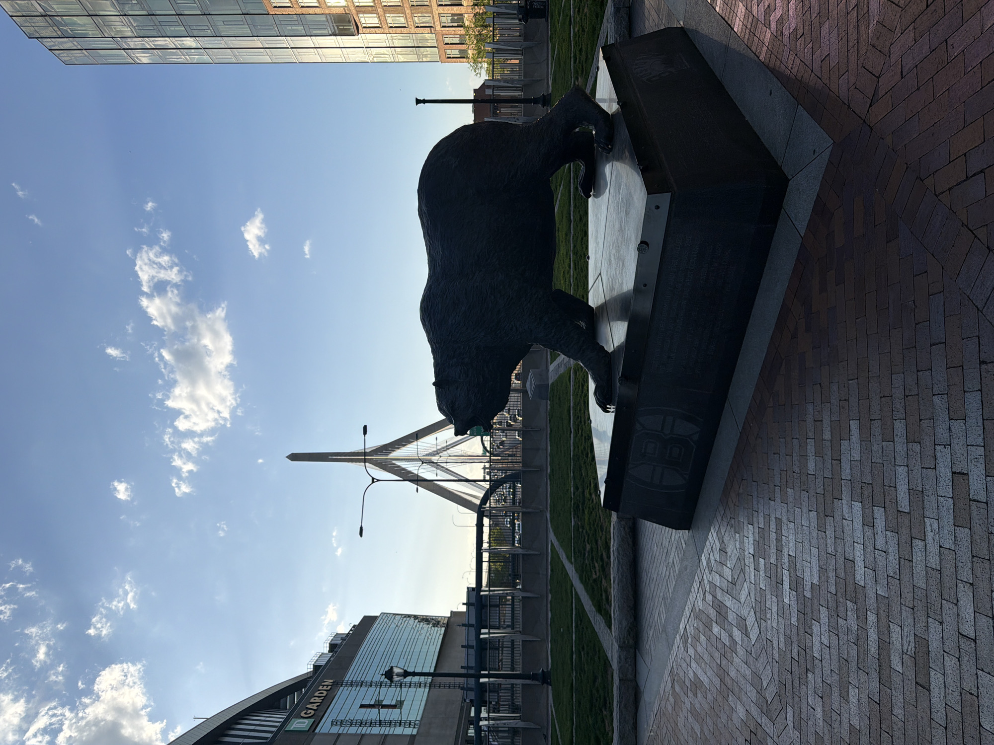 Here's a detailed description of the image:
**Overview:** The image features a bronze bear statue mounted on a dark, geometric stone base. Behind the statue, there's a view of the Zakim Bridge, a cable-stayed bridge in Boston, Massachusetts.
**Objects and Structures:**
* **Bear Statue:** The bear is depicted in a dynamic pose, likely representative of the Boston Bruins ice hockey team.
* **Statue Base:** The base appears to be made of dark stone.
* **Zakim Bridge:** The bridge is visible in the background, identifiable by its distinctive cable-stayed design.
* **TD Garden:** Part of the TD Garden arena is visible in the foreground, its name displayed on the building.
* **Buildings:** In the background, various buildings can be seen, some with modern architectural design.
* **Ground:** There is a brick or stone paved area in the lower right and an area of grass beyond the statue.
**Environment:** The scene appears to be set outdoors, possibly in a plaza or park setting near the TD Garden and the Zakim Bridge in Boston. The lighting suggests it's daytime.
**Location:** Based on the Zakim Bridge and the TD Garden, the image is located in Boston, Massachusetts, USA, near the arena and potentially in a related plaza or park area.
