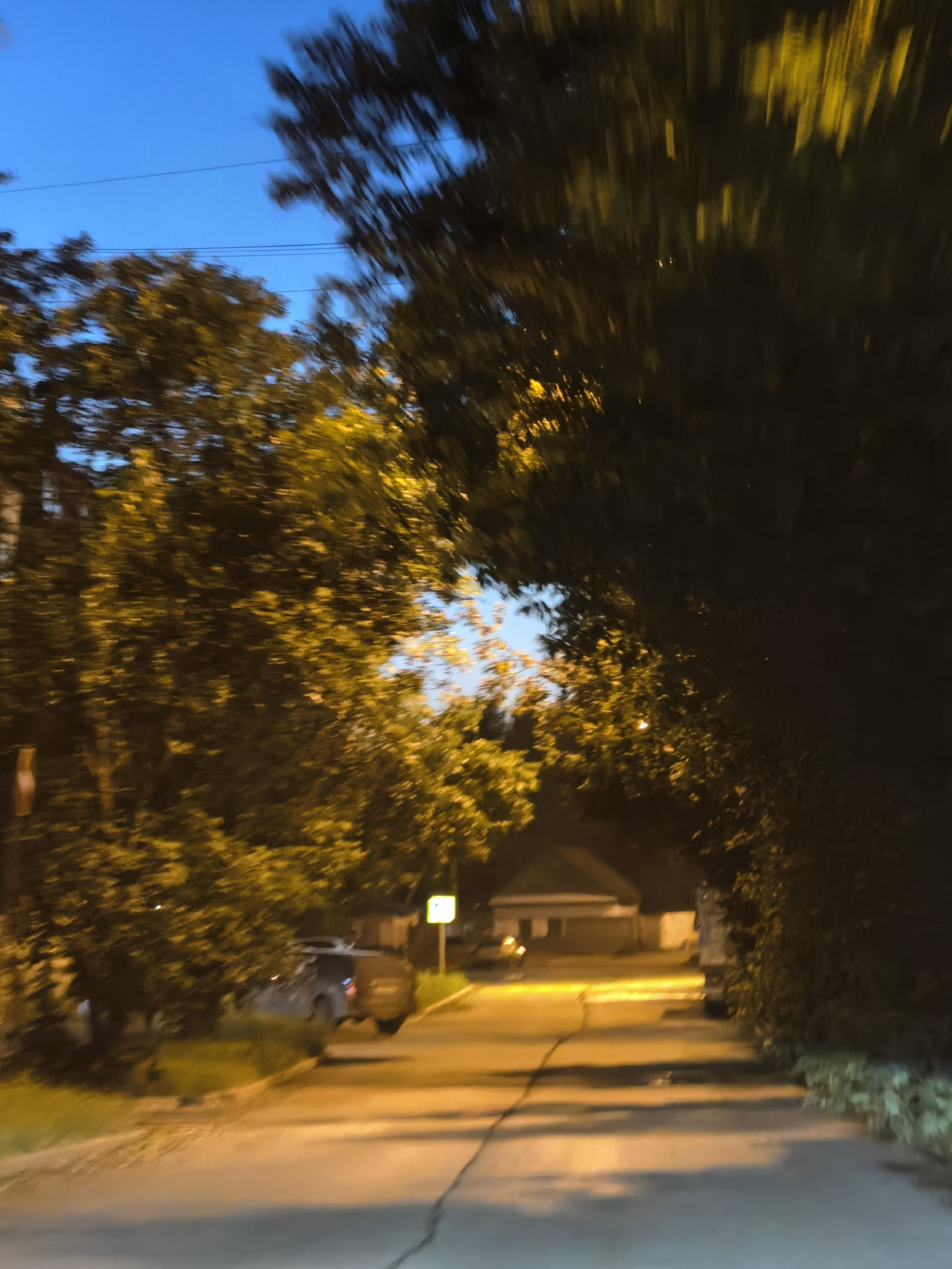 The image depicts a blurred view of a residential street at dusk. Trees line both sides of the road, their leaves illuminated by streetlights, casting a warm glow. The sky is a deep blue, suggesting twilight. In the middle ground, a car is parked on the side of the road. A house with a visible roofline can be seen in the distance. The overall impression is one of a quiet, suburban evening.