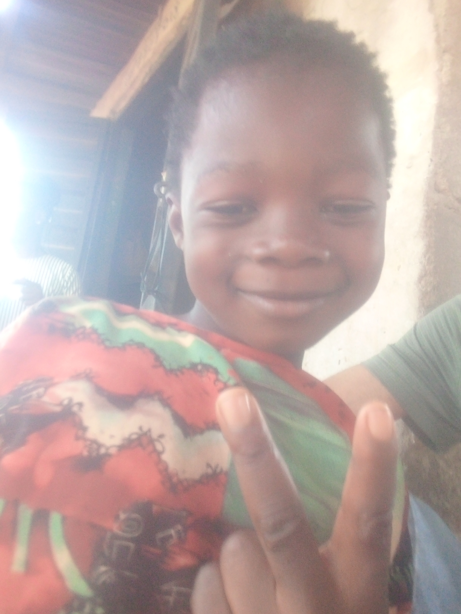 This media file captures a close-up, slightly blurry portrait of a young child with a dark complexion, smiling cheerfully at the camera. The child has short, dark, and curly hair. Their eyes are narrowed in a happy expression, and a wide smile is visible.
The child is wearing a vibrant, patterned garment, possibly a wrap or top, which features bold red, green, and black designs. With their right hand, they are making a peace sign (two fingers raised) towards the viewer, enhancing the impression of a playful and positive mood.
The background of the scene is somewhat indistinct due to blur and strong lighting. A prominent light source in the upper left creates a significant glare across that portion of the image. Above the child, wooden beams are visible, suggesting an indoor setting with a wooden ceiling or roof structure. To the left, behind the child, a person wearing a striped shirt is partially visible, and on the right side of the frame, a segment of a green garment, possibly an arm or shoulder, is also visible. The lighting is bright, but results in some overexposure and softness throughout the image. There is no visible text. The overall emotion conveyed is one of happiness and cheerfulness.