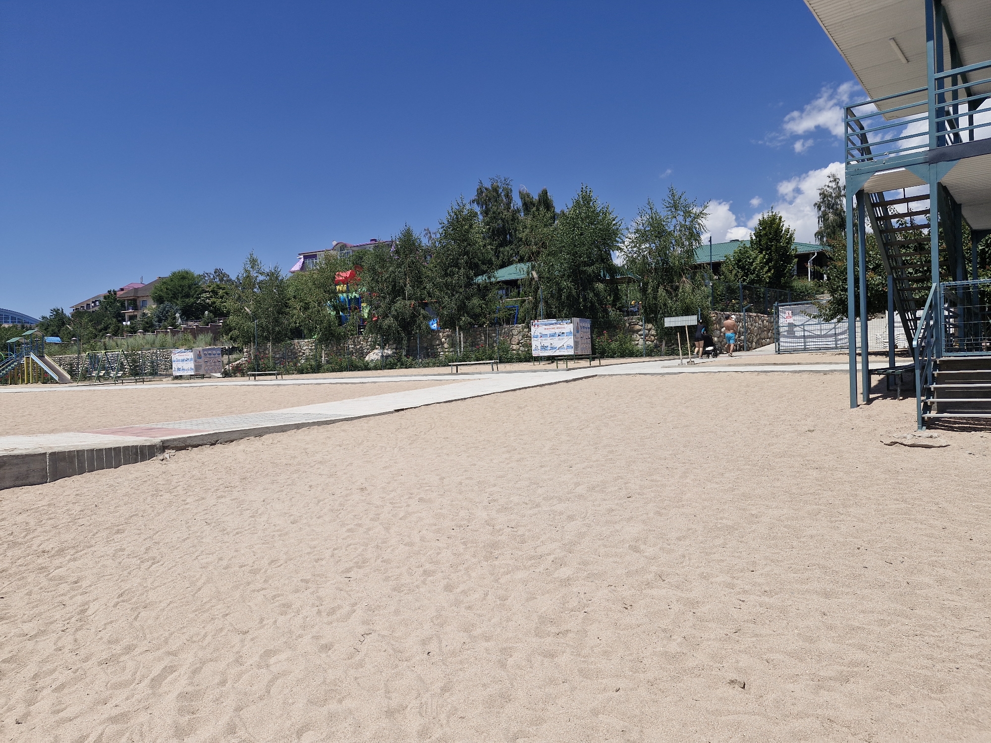 This is a wide-angle, eye-level photograph of a sandy beach area in Bulan-Segettu, Kyrgyzstan, under a clear blue sky with scattered clouds. The foreground is dominated by a vast expanse of light brown sand, showing slight undulations and a few scattered natural debris. A light-colored paved walkway with a reddish stripe runs diagonally from the lower left towards the upper right, bordered by a concrete curb. Beyond the walkway, in the middle ground, is a low stone wall and a fence line, behind which are trees and buildings. The trees are lush green, and some buildings have colorful roofs. On the left side of the middle ground, a playground structure with slides and swings is partially visible. Several park benches are placed along the walkway. On the right side of the image, a two-story lifeguard or observation tower with a metal structure, blue railings, and a white roof stands prominently, featuring an open staircase on its right side. Two individuals are visible in the middle ground: one person in blue shorts and another in dark clothing, standing near the fence. The overall scene depicts a well-maintained recreational area on a sunny day.