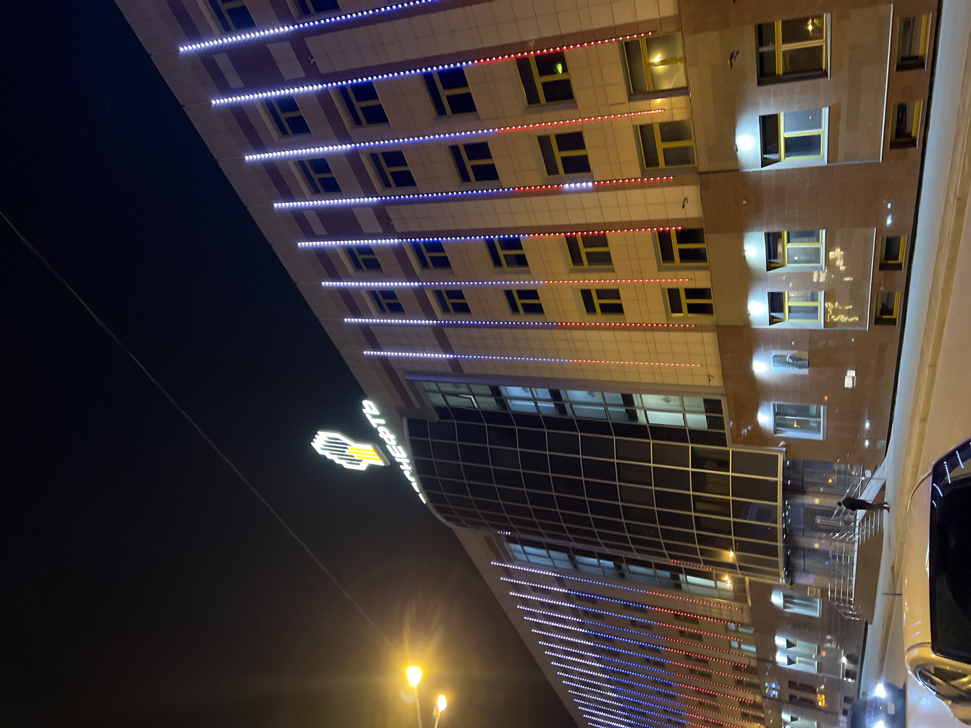 At night, the image depicts a tall, modern building in Tyumen, Russia. The building is illuminated with horizontal rows of LED lights, alternating between red, white, and blue. The upper section of the building features rectangular windows with yellow frames, set into a beige and brown facade. Below this, a large glass section is visible, reflecting the surrounding light. A sign with the name "Surgutneftegas" in Cyrillic script, along with a distinctive logo, is illuminated at the corner of the building. A street lamp with two lights shines in the foreground. A car is parked on the side of the building. A person walks along a walkway.