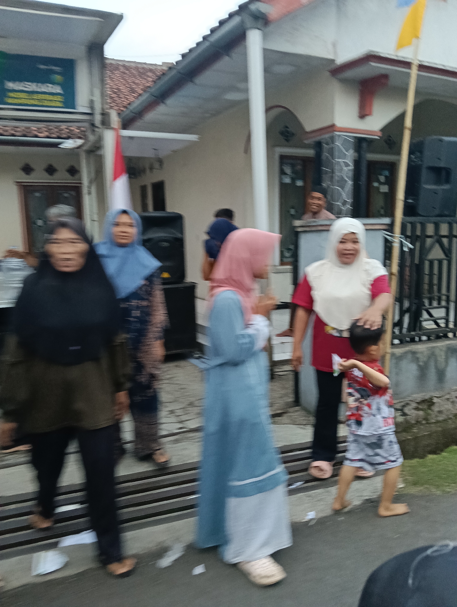 The image captures an outdoor scene in the city of Tasikmalaya, Indonesia, likely during an event or gathering. Several individuals, including women and a young child, are visible. One woman, wearing a black hijab and dark clothing, stands on the left. To her right, another woman in a light blue hijab and patterned dress is also present. Further to the right, a woman in a white hijab and red shirt is interacting with a young boy in a patterned shirt and shorts. The background shows a building with a tiled roof and a covered porch, as well as a speaker on a stand. A flag, possibly red and white, is also visible, suggesting a national or celebratory context. The ground appears to be paved, with some grass and a small ditch or drain visible on the right. The overall impression is of a casual, community-oriented event.