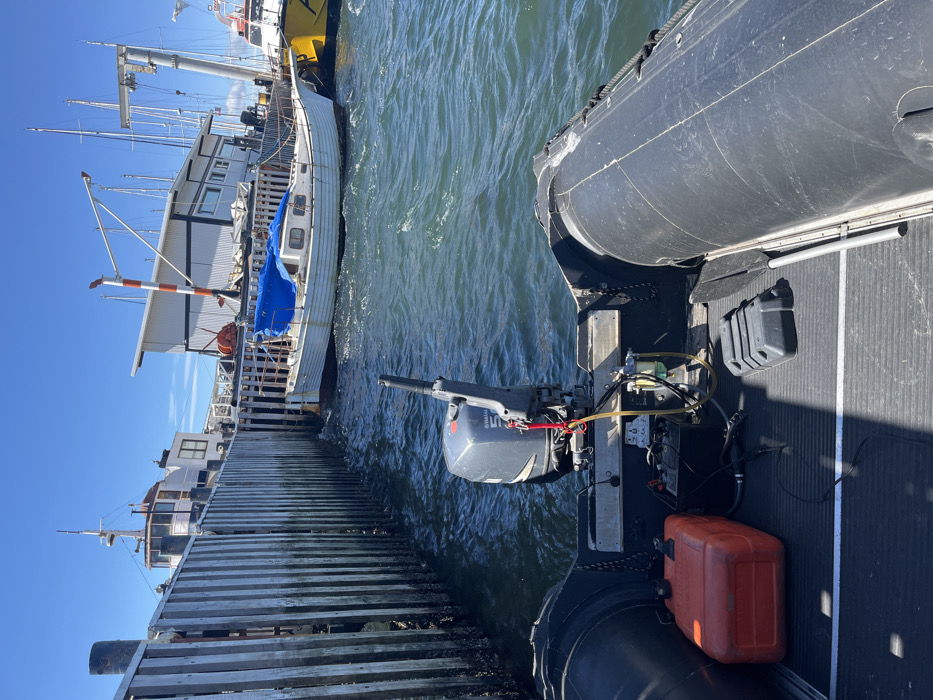 A black inflatable boat with an outboard motor sits alongside a wooden dock. The water is choppy and reflects the sunlight. On the other side of the dock, a larger white and yellow boat is docked. A blue tarp is draped over part of the white boat. Several masts are visible in the background. The sky is clear and blue. An orange fuel canister sits on the floor of the inflatable boat.