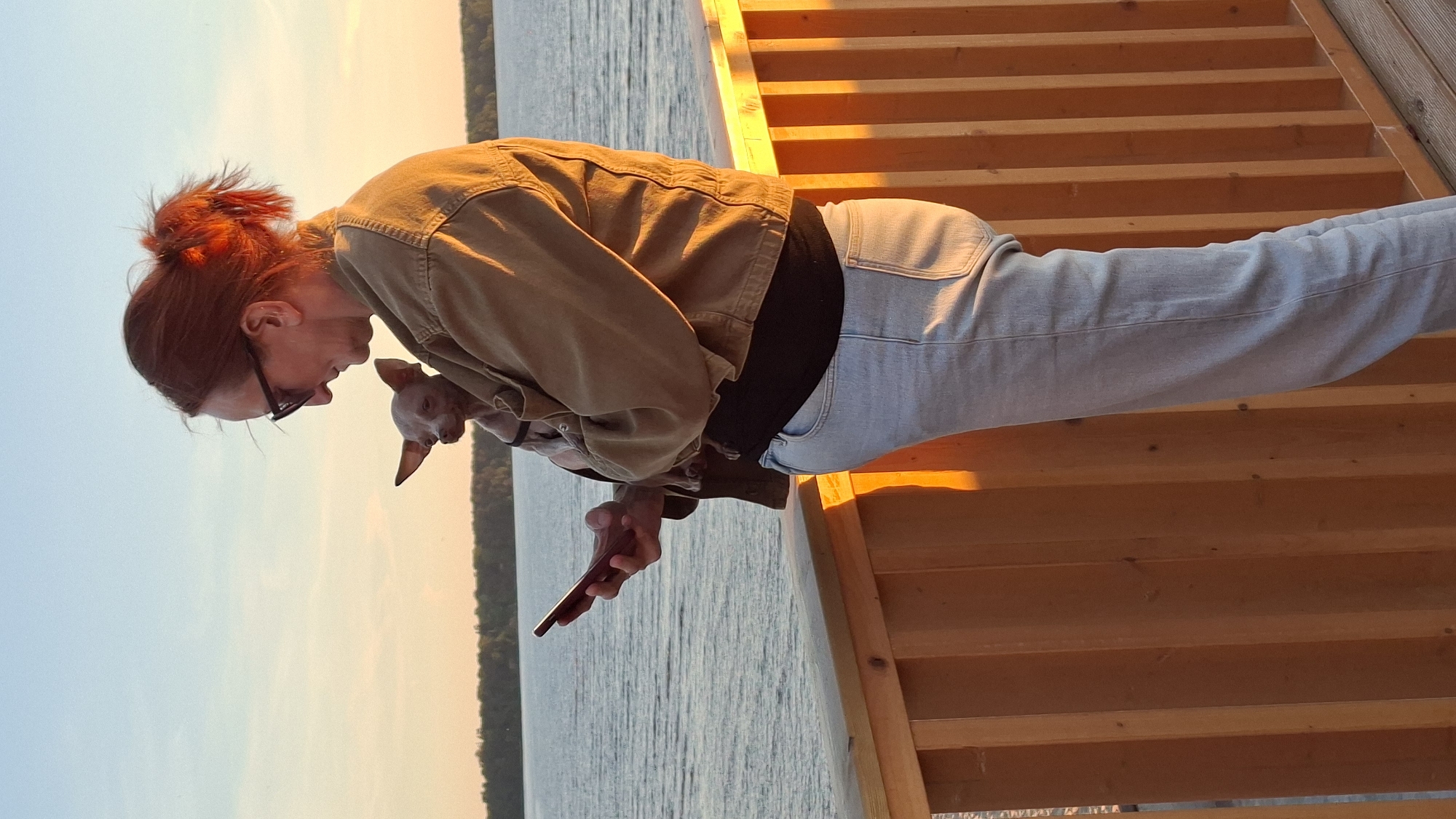 A woman with auburn hair pulled into a messy bun and wearing glasses stands on a wooden deck, holding a small, light-colored dog in her arms. She is looking down at her smartphone. The woman is wearing a khaki-colored jacket and light-wash jeans. Behind her is a body of water, likely a lake or the sea, with a tree line visible in the distance. The sky is a pale blue with hints of orange from what appears to be a sunset. The wooden railing of the deck is in the foreground, and the setting sun casts a warm glow on the scene. The location is the city of Tallinn, Estonia.