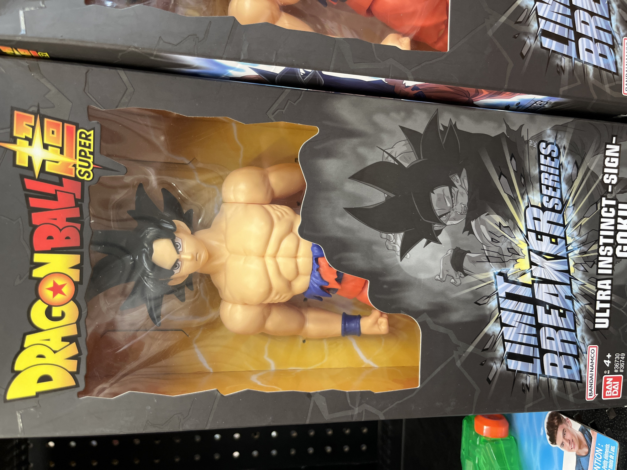 The image shows a close-up of a Dragon Ball Super action figure in its packaging. The figure is Ultra Instinct -Sign- Goku, and the packaging is part of the "Limit Breaker Series". The packaging is black with a yellow and red "DRAGON BALL SUPER" logo on the left. The figure itself is visible through a plastic window, and the box also features a black and white illustration of Goku in the Ultra Instinct form. The age recommendation is 4+. The toy is located on a store shelf.