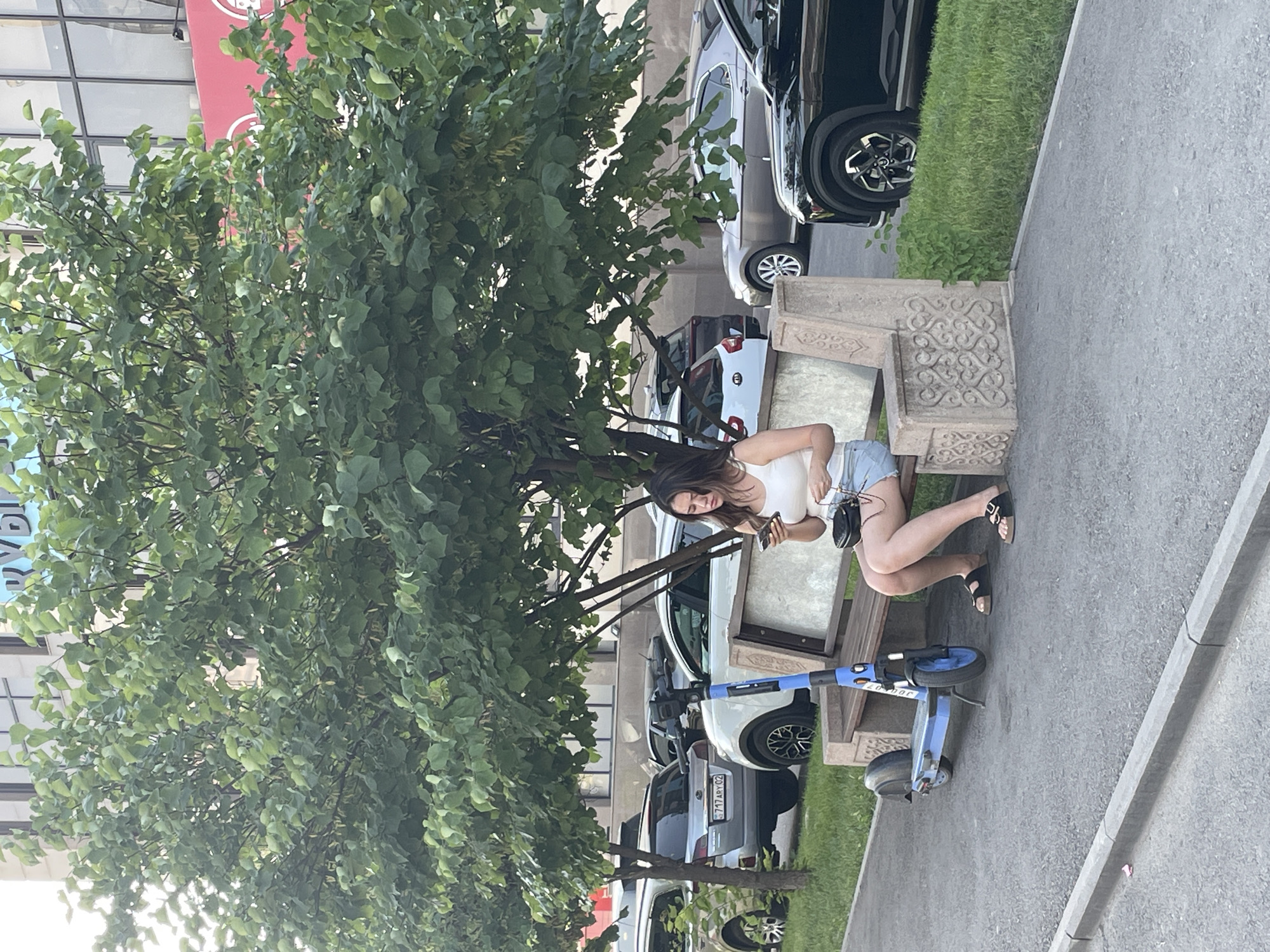 A young woman is sitting on a decorative concrete bench under a large tree, using her phone. She is wearing a white tank top and denim shorts. A blue electric scooter is parked next to the bench. In the background, cars are parked along a street, and a building with red and white signage is visible. The scene appears to be in a city, likely Almaty, Kazakhstan, given the context.