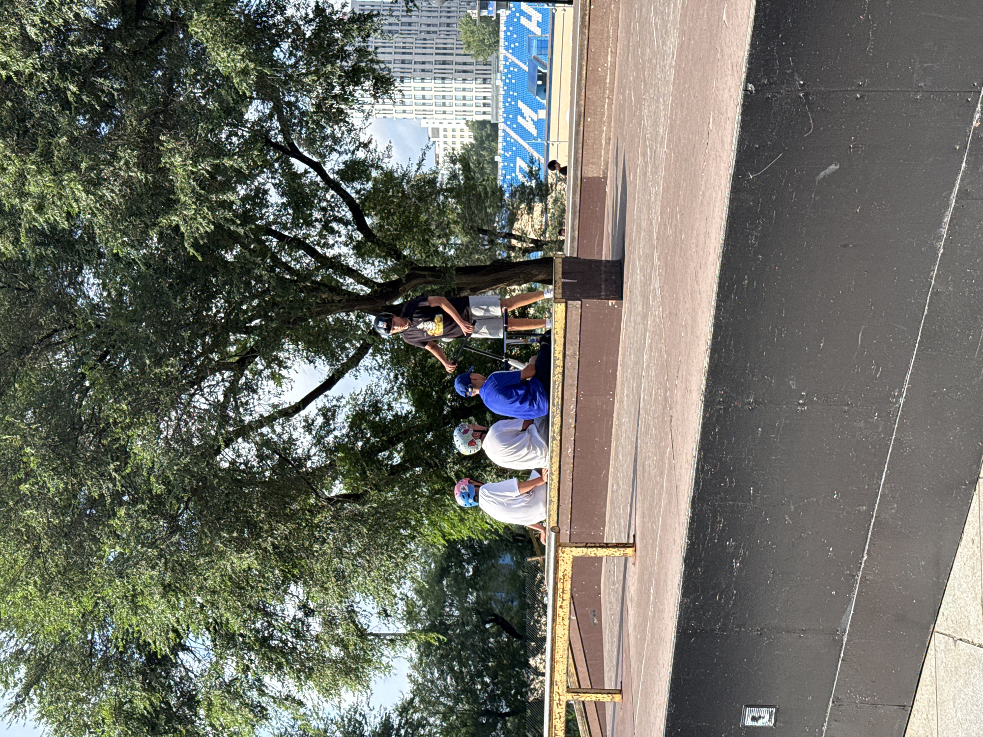 Four youths are gathered at a skate park in Vladivostok, Russia. Three of them, wearing helmets, are seated on a ramp. One youth, also wearing a helmet, stands with a scooter. In the background, a stadium with blue seating is visible, along with tall buildings. The scene takes place outdoors under a large tree on a sunny day.