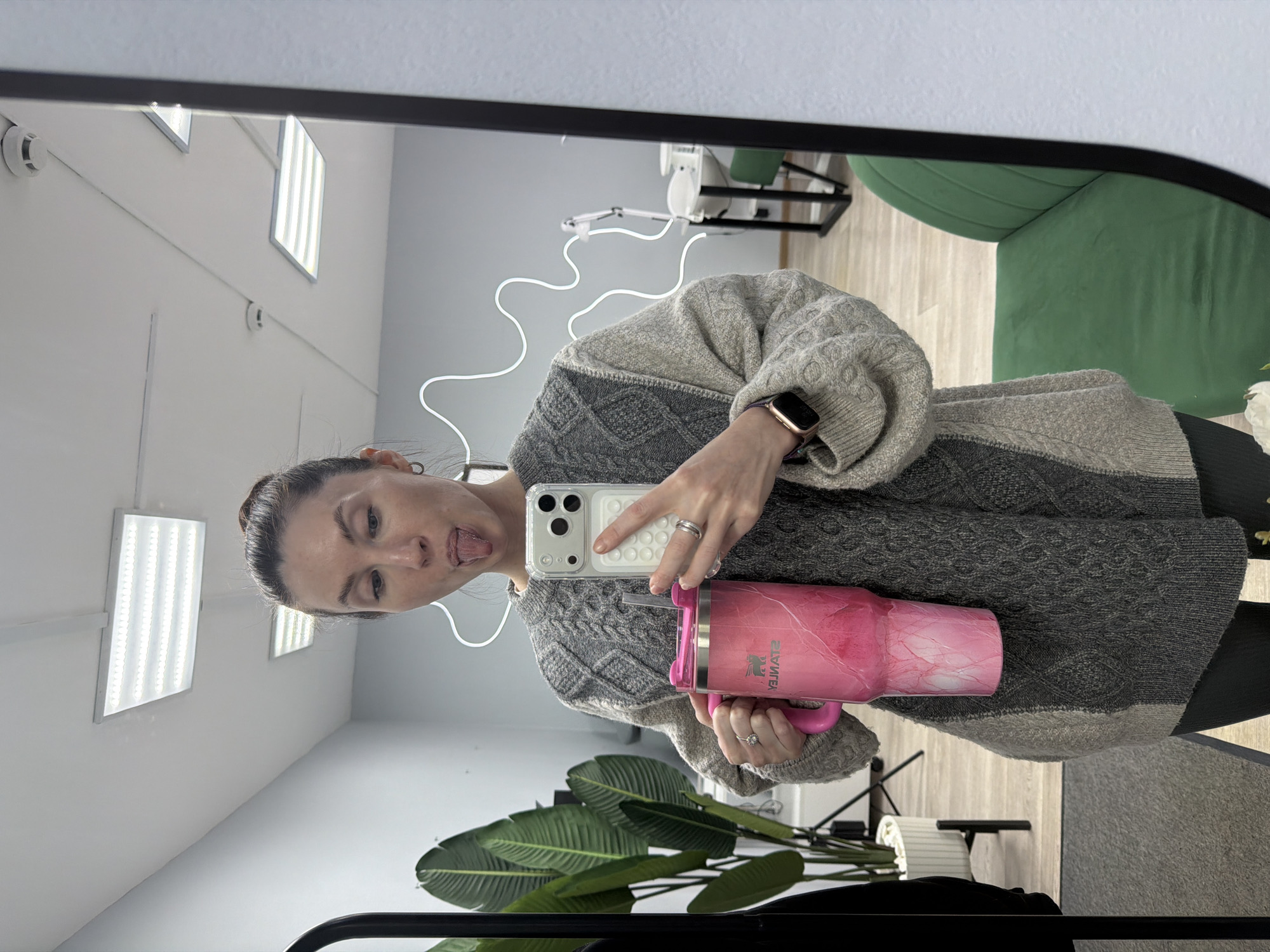 A Caucasian woman with her hair in a bun takes a selfie in a mirror. She is wearing a gray and beige sweater. She holds a pink Stanley tumbler in her left hand and a cell phone in her right. The background shows a white wall with rectangular lights, a green chair, and a plant. The image was taken in an enclosed setting, likely a room. The location in Perm, Russia is not discernible.