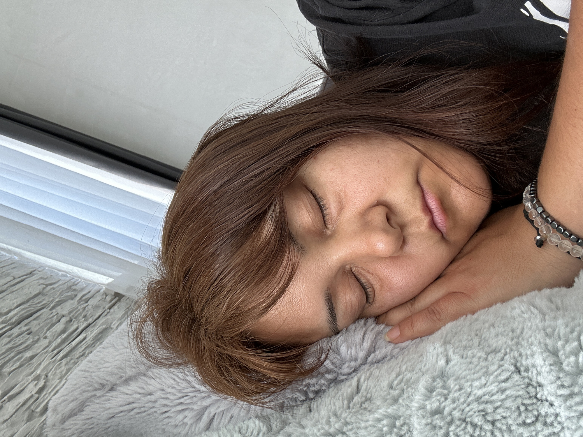 A young adult woman is lying down on a grey, fluffy surface with her eyes closed. Her head is resting on her left hand, which is adorned with a beaded bracelet. She has brown hair with highlights and a fair complexion. She is wearing a black shirt. In the background, there's a window with white blinds and a white wall. The location is Unknown, Unknown.
