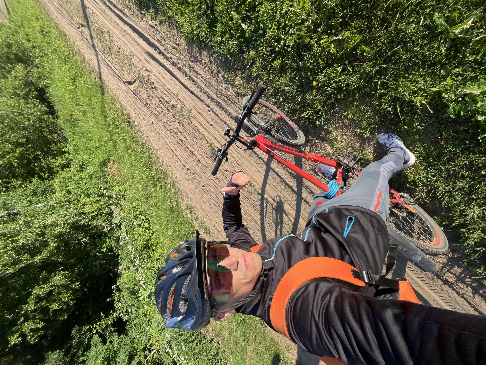 A man wearing a black cycling jacket, grey pants, a black helmet with orange accents, and reflective sunglasses is taking a selfie while sitting on a red bicycle on a dirt road in Chaikovsky, Russia. The road is surrounded by green grass and trees, suggesting an outdoor, natural environment. The man is giving a thumbs-up gesture. The bicycle has black handlebars and red frame. The lighting suggests it is daytime.