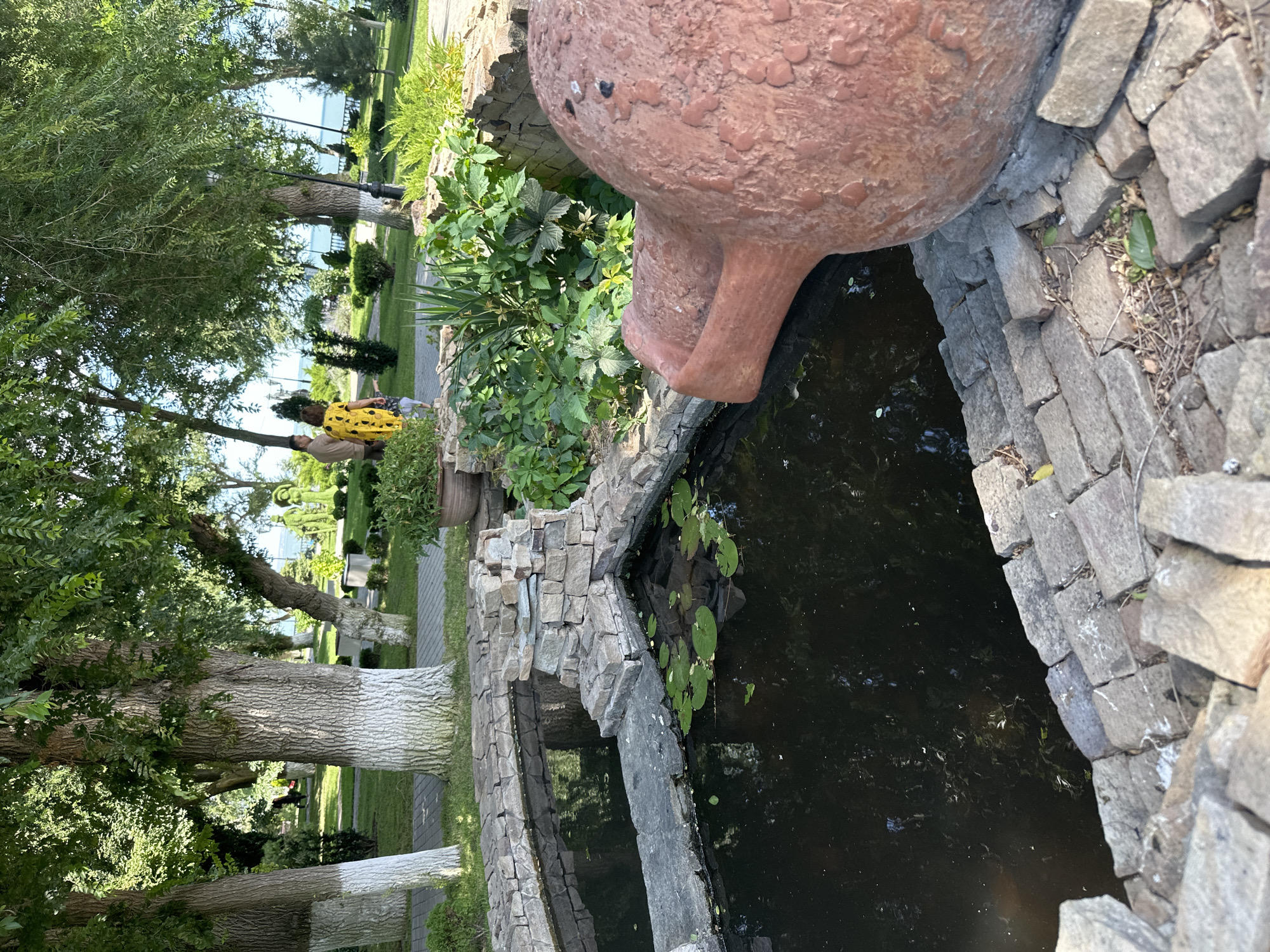The image depicts a park area with a decorative pond in the foreground. The pond is constructed with rough-hewn stones and contains dark water with lily pads. To the right of the pond is a large, reddish-brown decorative pot or sculpture. Behind the pond, a paved walkway leads through a grassy area with trees. In the mid-ground, two people are walking, one wearing a yellow dress with a black pattern. Beyond the trees, a body of water, likely the sea, is visible, suggesting the park is located near the coast. The overall environment is lush and green, indicative of a park or garden setting in Taman, Russia.