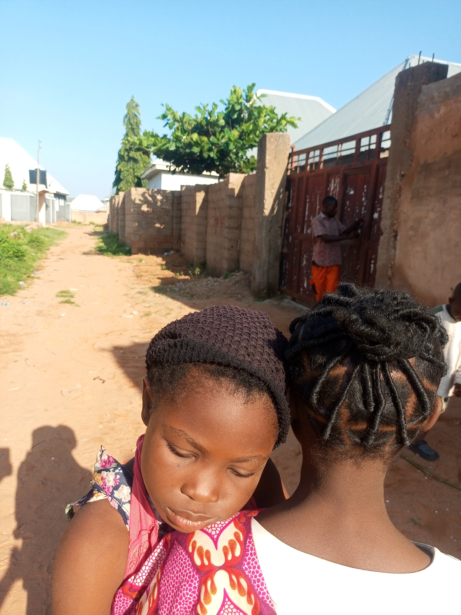 In Tempure, Nigeria, an over-the-shoulder view captures two young girls in the foreground, with a man visible in the background near a gate. The girl in the left foreground, closer to the camera, wears a dark, textured knit cap and a vibrantly patterned dress with floral and abstract designs, a pink scarf draped around her neck. Her eyes are closed, and her head is bowed. To her right, another girl's head is visible from behind, her dark hair styled in intricate braids. Behind them, a dirt path stretches into the distance, bordered by a low, sandy brick wall on the right and buildings on the left, including one with a silver corrugated metal roof and another with a white facade. A large, reddish-brown metal gate stands partially open, with a man in a light-colored, short-sleeved shirt and orange trousers standing near it, appearing to interact with the gate. The sky above is a clear, bright blue.