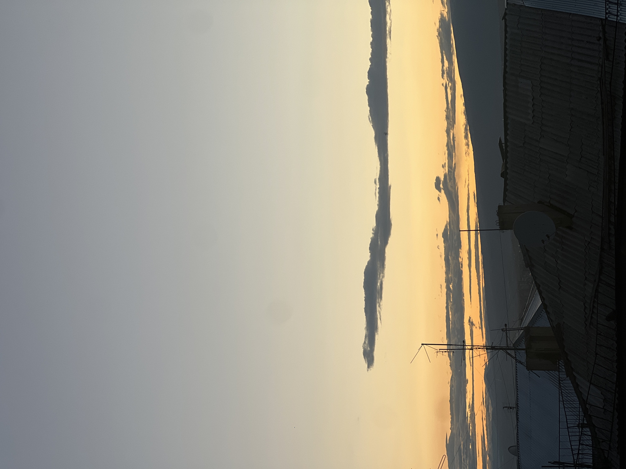 The image shows a portion of the sky at sunset, with shades of orange and yellow. A thin, dark cloud stretches vertically across the sky. Part of a building is visible on the right, with a corrugated metal roof and a satellite dish. There is an antenna mounted atop the building. The image was probably taken in Yerevan, Armenia.