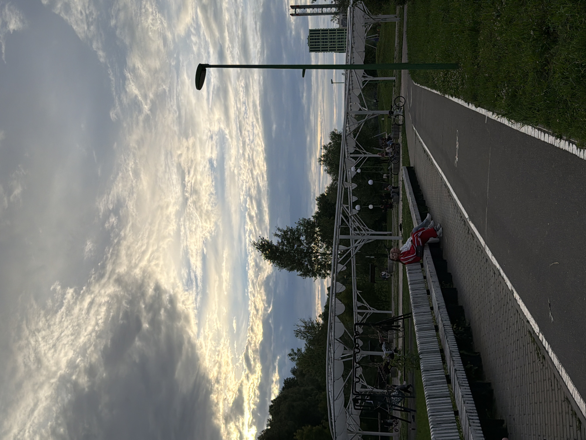 The image depicts a park scene in Kazan, Russia, with a cloudy sky and trees in the background. In the foreground, a paved path runs alongside a long bench where two individuals are seated, wearing red and white athletic wear. Beyond the bench, a white canopy structure is visible, resembling a gazebo or a covered walkway, with playground equipment underneath. A tall green lamppost stands to the right of the path. In the distance, two modern buildings, one green and one with red structural elements, are visible, suggesting an urban edge to the park. The overall atmosphere is calm, with people enjoying a leisurely time in the park.