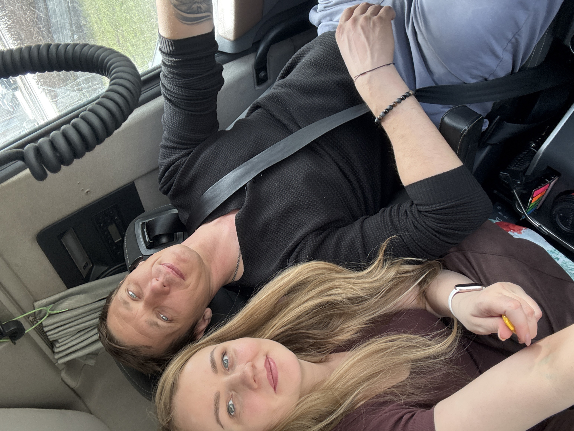 Two people, a man and a woman, are inside what appears to be a vehicle. The man is wearing a black long-sleeved shirt and is wearing a seatbelt. The woman has long blonde hair and is wearing a dark brown shirt and a smart watch. The interior of the vehicle is visible, including a coiled cable. The location is not discernible as Izhevsk, Russia from the image.