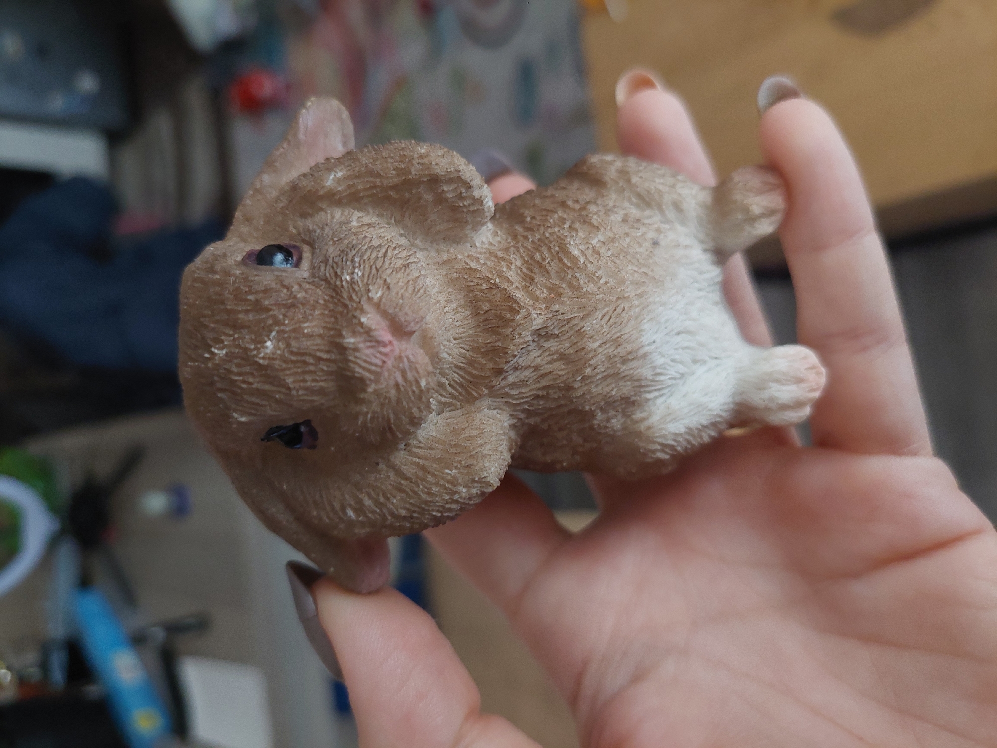 A person holds a small, brown, textured rabbit figurine in their left hand. The figurine is lying on its side. The background shows a blurred interior scene with various objects. It's impossible to determine whether this is in Plovdiv, Bulgaria, or not, given the limited information.