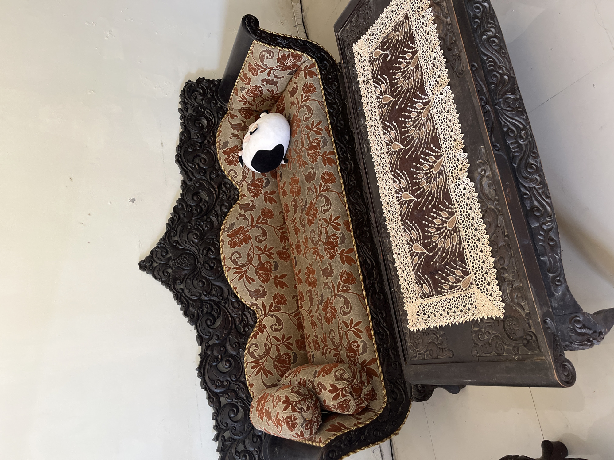 The image shows an ornate, dark brown wooden sofa with intricate carvings on its frame. The sofa is upholstered in a fabric with a floral pattern in shades of brown and red. A round, stuffed toy with black and white markings is placed on the right side of the sofa. In front of the sofa is a matching dark brown wooden coffee table. The table is also ornately carved and has a decorative lace runner with a brown background and white lace trim, featuring a peacock feather-like design. The floor appears to be tiled in a light color. The background consists of a plain, off-white wall. The scene is set indoors, likely in a living room. No specific Sidoarjo landmarks are visible, but the style of the furniture could be indicative of Indonesian craftsmanship.