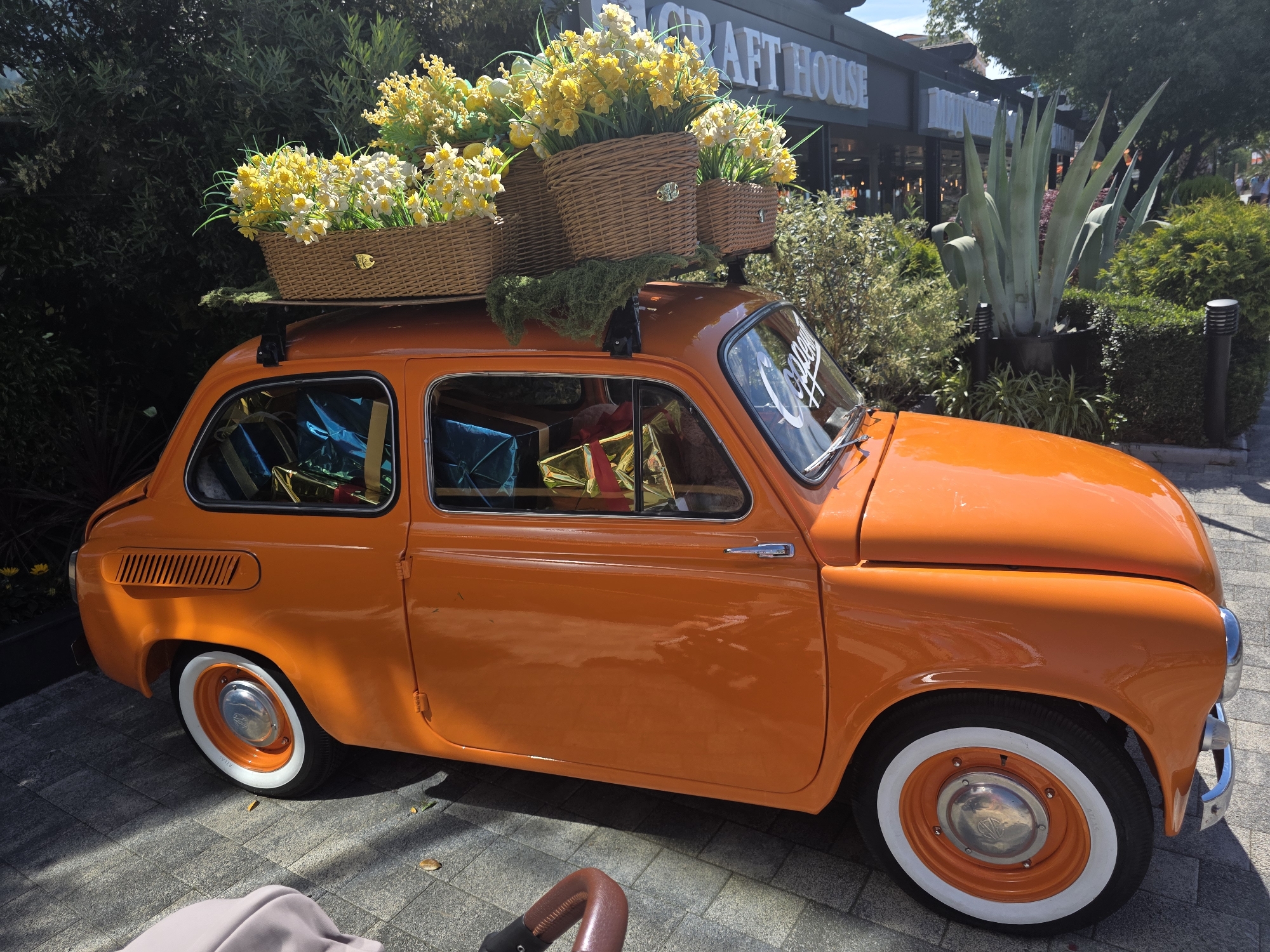 An orange vintage car, possibly a modified Fiat 600 or similar microcar, is parked on a light grey paver stone surface outdoors in Sochi, Russia. The vehicle is brightly polished, reflecting direct sunlight, and features white sidewall tires with orange-painted wheel hubs and chrome hubcaps. A roof rack supports three wicker baskets overflowing with artificial yellow and white daffodil-like flowers, with green moss-like material draped around the bases of the baskets. Through the car's windows, several wrapped gifts in metallic blue and gold paper are visible inside the cabin. A white decal, partially legible as "Coopers" or similar, is affixed to the passenger side of the windshield. In the background, a dark-colored building with prominent white block lettering spelling "CRAFT HOUSE" is visible, surrounded by lush green foliage, including various shrubs and a large agave plant in a planter. The scene is illuminated by strong daylight, casting defined shadows. A brown leather-like handle and light-colored fabric, likely from a stroller, are partially visible in the bottom left foreground.