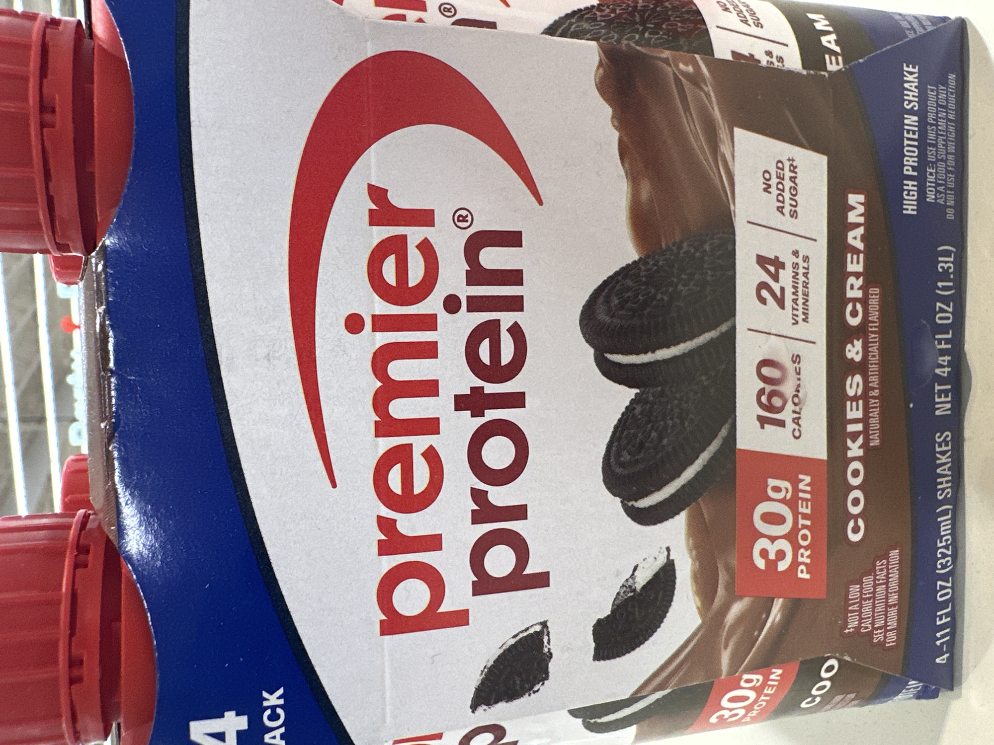 The image shows a close-up of a carton of Premier Protein Cookies & Cream flavored shakes. The carton features the brand's logo, a red arch over the words "premier protein". Below the logo, there are graphics of chocolate shakes and Oreo cookies. Text on the carton indicates that each shake contains 30g of protein, 160 calories, and 24 vitamins and minerals. It also states "No Added Sugar". The bottom of the carton shows "4-11 FL OZ (325mL) SHAKES NET 44 FL OZ (1.3L)". The product is identified as a "HIGH PROTEIN SHAKE" and includes a notice to use it as a food supplement only and not for weight reduction. The image appears to be taken in a retail environment, likely a grocery store, given the background lighting which suggests fluorescent lighting commonly found in such stores. The specific location of Camp Hill, United States of America is not explicitly visible in the image, nor are there any people or actions depicted.