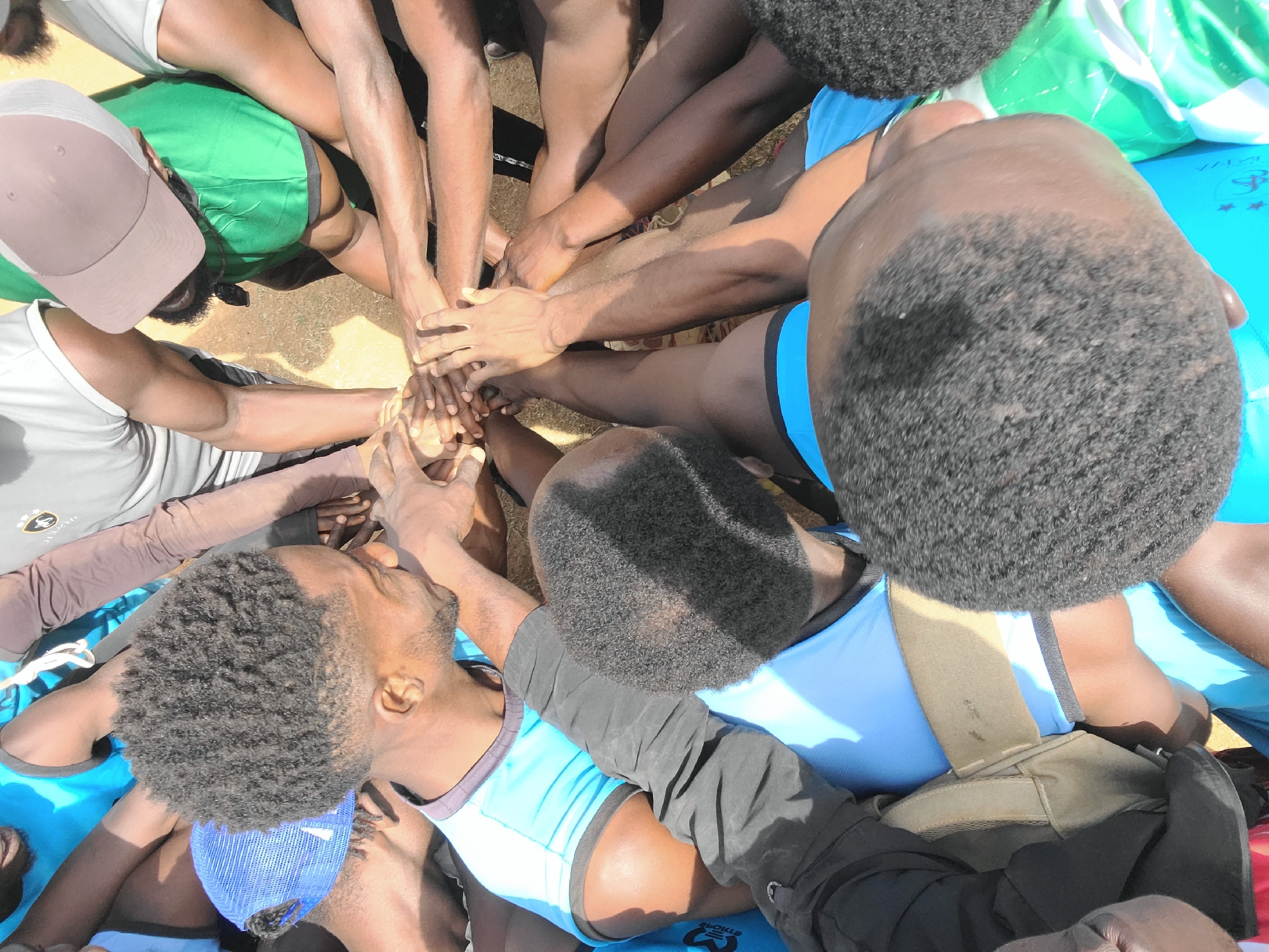 This close-up, high-angle image depicts a group of young men in Barikin Jirga Summa, Nigeria, gathered in a tight huddle with their hands stacked in a central pile, symbolizing unity and teamwork. Approximately six to eight individuals are visible, their dark skin tones contrasting with various athletic wear.
The men are dressed in a mix of sportswear: a light grey t-shirt with green shorts and a pink baseball cap is visible on one participant, while others wear light blue tank tops (some with dark trim), a white t-shirt, and dark long-sleeved garments. One man in the foreground wears a blue mesh baseball cap. Their short, curly hair is also a prominent feature.
The setting appears to be outdoors on a bright, sunny day, evidenced by the strong shadows and highlights. The ground visible beneath and around them is a light, dry color, suggesting sand or dirt. The activity conveys a sense of camaraderie, focus, and collective spirit, possibly preceding a sporting event or a group endeavor.
Notable details include faint visible text and logos on some items. On a light blue fabric or mat, white text that might read "VALLEY" along with a star-like emblem is partially discernable. Another small logo with letters resembling "ST" is faintly visible on a dark blue garment. The primary focus of the image is on the interconnected hands and arms, emphasizing the group's cohesion.