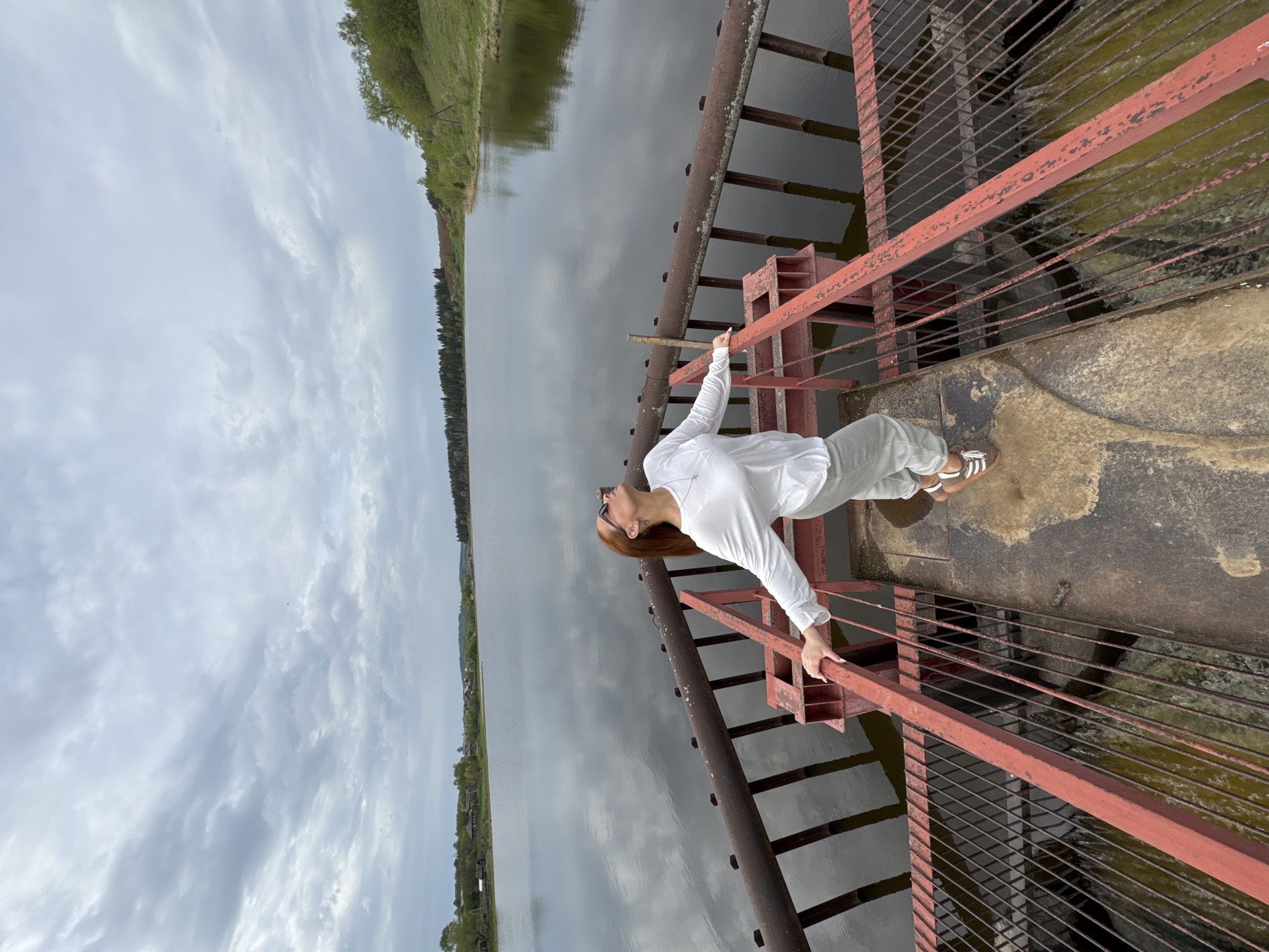 A woman wearing a white long-sleeved shirt and light-colored pants stands on a rusty red metal structure, possibly a bridge or dam, with a body of water visible below and in the background. She is leaning back, holding onto the railing with both hands, and looking upwards. The water reflects the cloudy sky. The opposite bank is lined with trees and some vegetation. The image suggests a rural or natural setting, possibly taken in or near the city of Nozhovka, Russia, given the prompt's specifications.