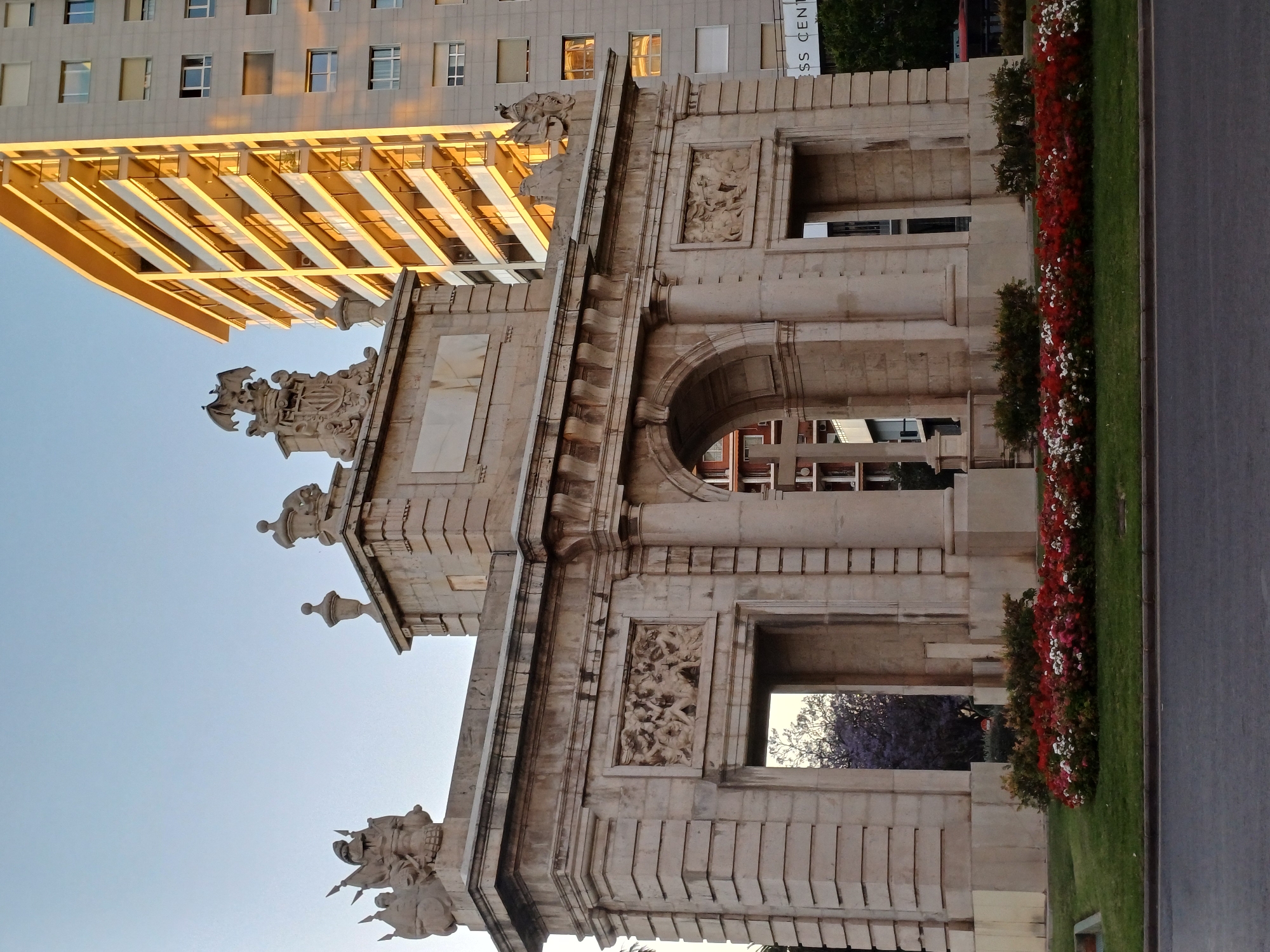 The image captures a close-up of a historical stone archway, possibly a gate or entrance, with intricate carvings and classical architectural details. Above the archway are sculptures and decorative elements. Adjacent to the archway is a manicured green lawn with flowerbeds featuring red and white blossoms. To the left, a modern, multi-story building with a pale facade and numerous windows rises behind the archway. The lighting suggests it is either early morning or late afternoon. The presence of the building juxtaposed with the historic architecture indicates an urban setting.
Given the characteristics in the image, the structure is likely one of the gates of the city of València, Spain.