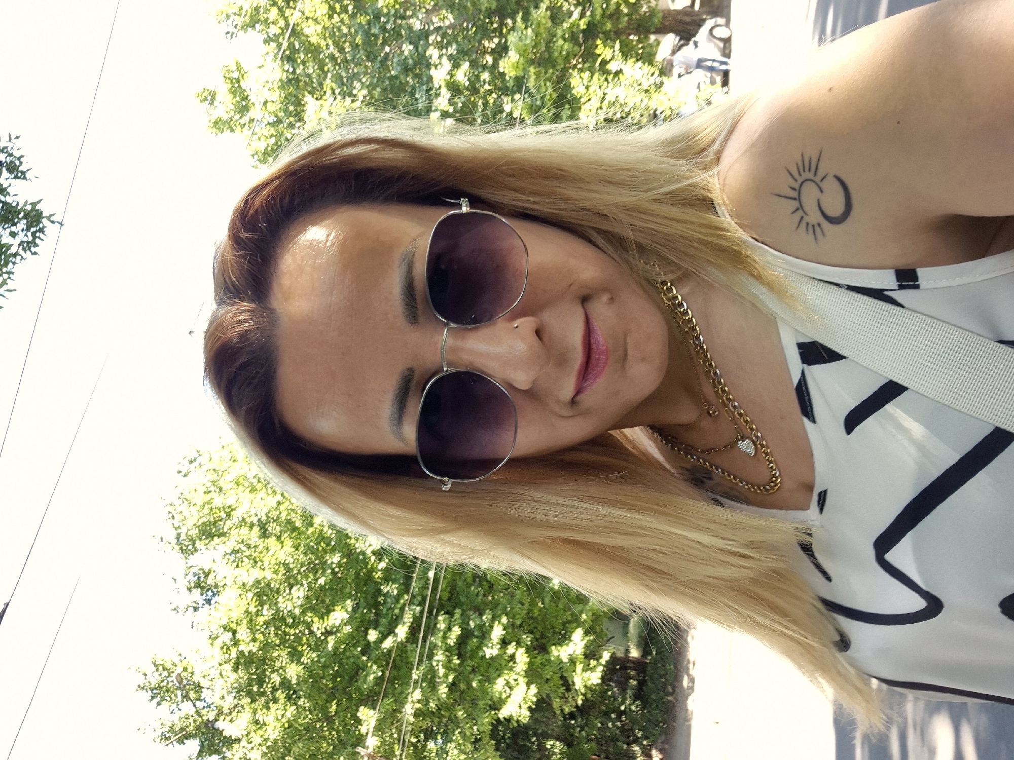 A woman is posing for a selfie outdoors. She has blonde hair and is wearing sunglasses, a necklace, and a white top with a black abstract pattern. She has a tattoo of a sun and crescent moon on her left shoulder. The background consists of trees, a sky with some power lines, and a blurred street with a car and a person in the distance. The lighting suggests it is daytime.
