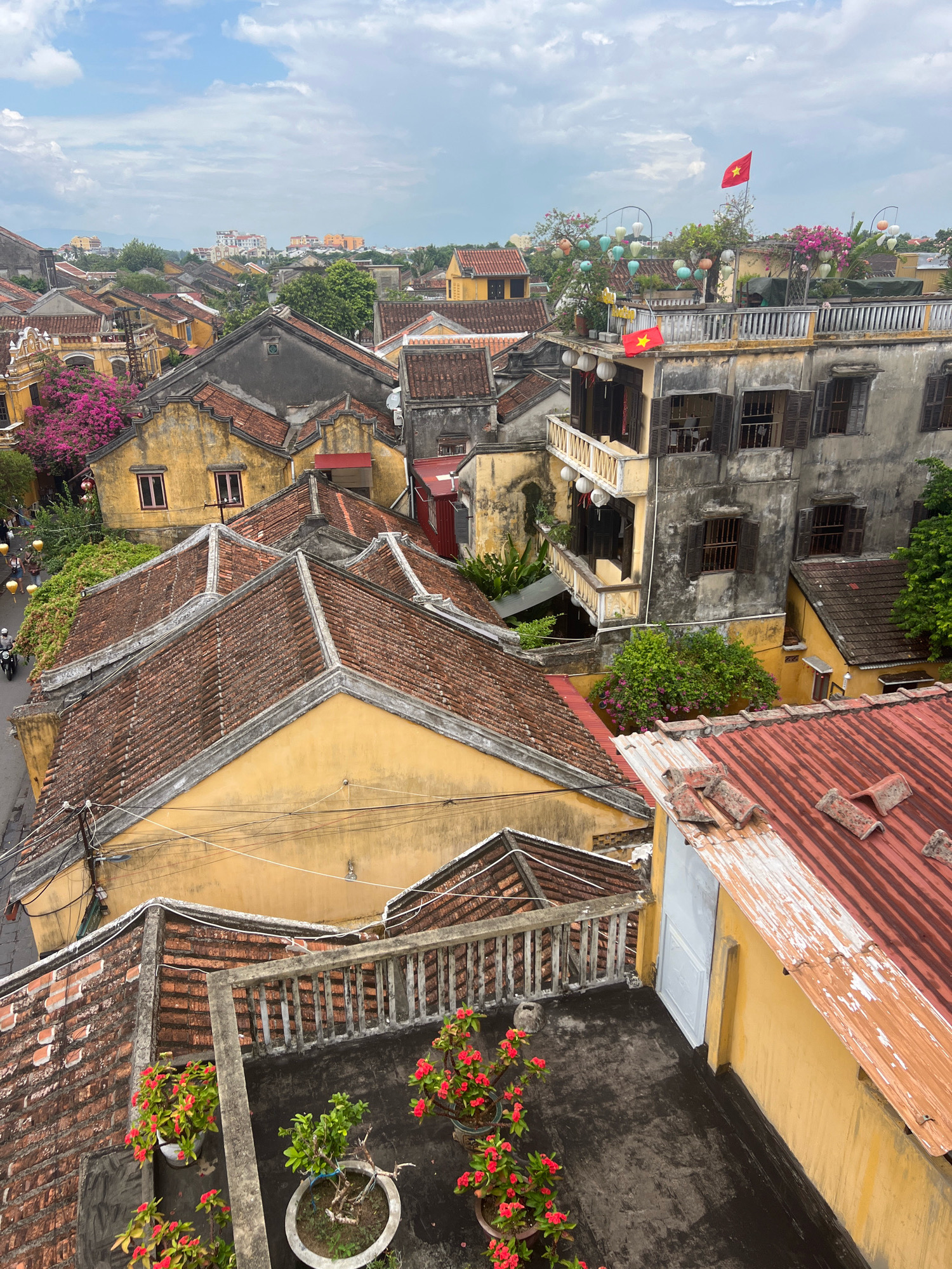 This is a high-angle, eye-level, medium shot of a rooftop view in the city of Hoi An, Vietnam. The scene is dominated by the characteristic yellow and grey architecture of the historic town, with many buildings featuring red tile roofs. In the foreground, a dark-colored rooftop area is visible with several potted plants, some bearing small red flowers. A concrete railing runs along the edge of this rooftop.
Further into the image, the dense urban landscape unfolds, with tightly packed houses and buildings. Several structures are painted in faded yellow, a common color in Hoi An, with contrasting red tile roofs. Some buildings exhibit weathered, grey facades. A balcony with decorative yellow railings extends from a grey building. Various trees and lush greenery are interspersed among the buildings, some with vibrant pink flowering trees.
In the distance, more buildings can be seen, and a red Vietnamese flag with a yellow star flutters from a rooftop. String lights and traditional lanterns are also visible, suggesting a festive or evening ambiance. On a street below, a few people and a motorbike can be observed, indicating the presence of daily life in the town. The sky is partly cloudy, with a mix of blue sky and grey clouds. The overall impression is a panoramic view of the charming, historic district of Hoi An.