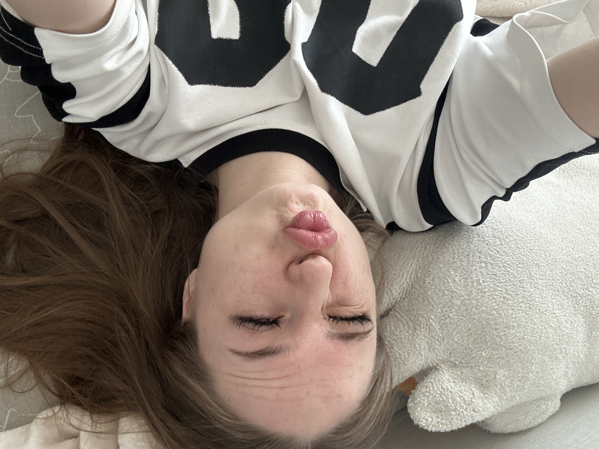 A person is depicted in a close-up selfie, lying down on their back. Their head is angled towards the camera, with long brown hair fanning out around them against a light-colored surface, possibly bedding or a blanket. They are wearing a white long-sleeved top featuring prominent black graphic elements, which appear to be large numbers, though not fully visible for identification. The individual's face displays a playful or silly expression, characterized by puckered lips in a "duck face" pose and eyes squeezed tightly shut, creating wrinkles on their forehead and around their eye area. A soft, white, textured object, likely a pillow or stuffed animal, is partially visible to the right of their head. The lighting is even and soft, suggesting an indoor setting. The image conveys a lighthearted and relaxed mood.