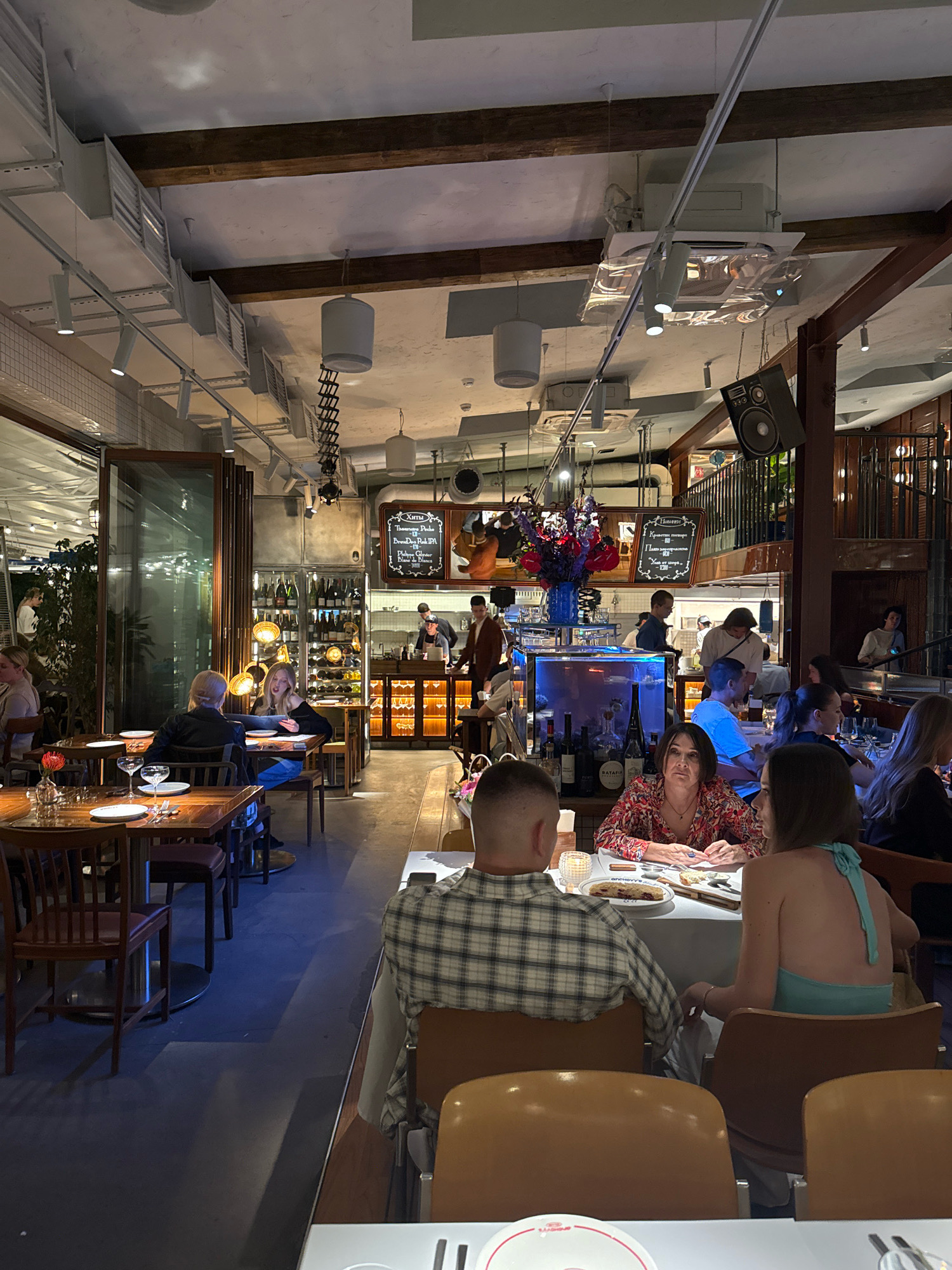The image depicts the interior of a restaurant in Moscow, Russia. The establishment features a modern industrial aesthetic with exposed wooden beams and ductwork on the ceiling. Lighting is provided by a combination of track lighting, pendant lamps, and accent lights highlighting a wine display and a bar area. Several tables are occupied by diners. In the foreground, a couple is seated at a table, with a woman in a patterned dress and a man in a plaid shirt. Further back, other patrons are seen dining and interacting. The bar area is visible in the background, with staff members preparing drinks. A prominent feature of the restaurant is a large glass aquarium containing fish, situated behind the bar. The overall ambiance suggests a lively and contemporary dining experience.
