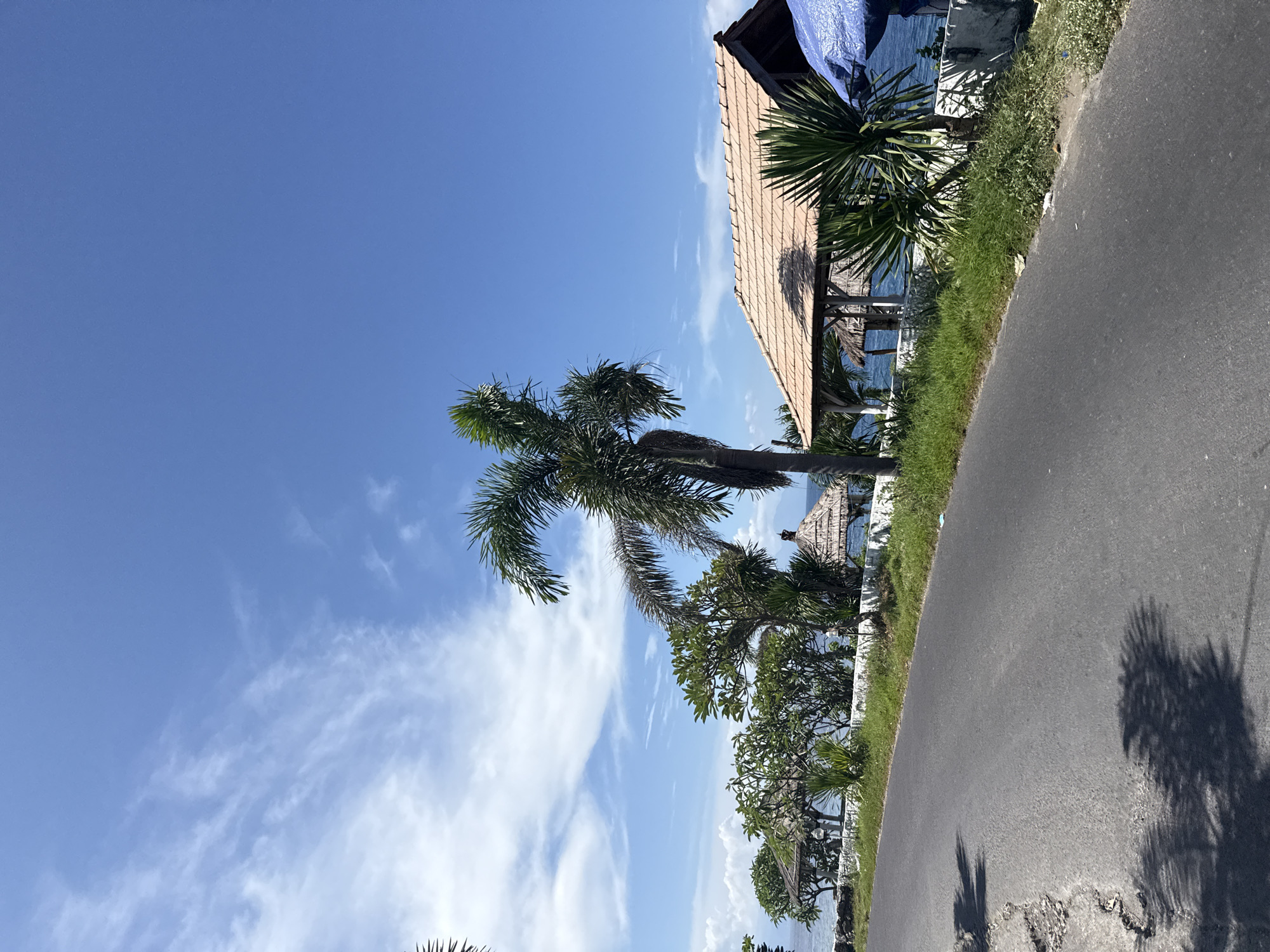A roadside scene in Batununggal, Indonesia is captured in this image. The foreground is dominated by an asphalt road, showing signs of wear. Along the road, there is a strip of greenery and some buildings with wooden roofs that border the sea. Palm trees are a prominent feature, standing tall. The sky is predominantly blue with streaks of white clouds.