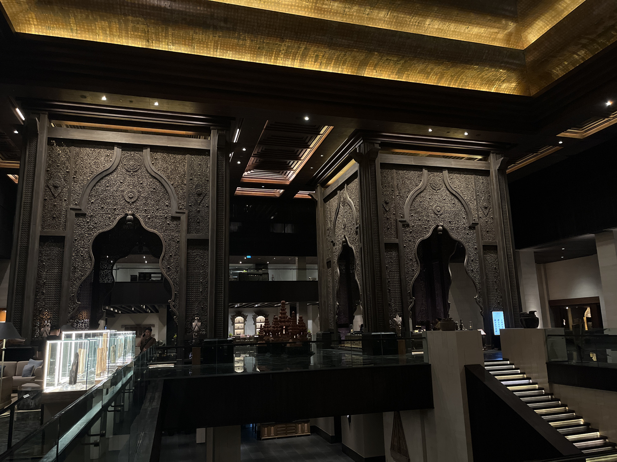 The image captures a grand interior scene, likely a hotel lobby or a similar opulent establishment, in Kuta Selatan, Indonesia. The architecture features towering, intricately carved dark wood structures that resemble ornate gateways or pillars, decorated with detailed patterns and arched openings. Above these structures, a luminous golden ceiling panel casts a warm, ambient glow throughout the space.
In the foreground, a modern staircase with illuminated steps ascends to the right, bordered by glass railings. To the left, a contemporary display case filled with various wooden objects is visible, with two individuals standing nearby. One person appears to be engaged in conversation on a mobile phone, leaning against a railing, while another is partially visible behind the display case, seemingly attending to it.
The midground reveals more of the expansive interior, with elevated walkways and additional decorative elements. A prominent, multi-tiered wooden sculpture sits on a reflective surface, suggesting a central artistic feature. Deeper within the space, glimpses of other seating areas and architectural details can be seen through arched openings. The overall impression is one of luxurious and culturally inspired design, illuminated by dramatic lighting.