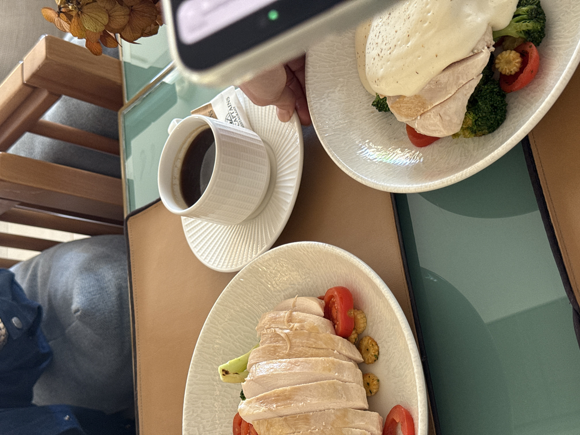 The image shows a table setting with two plates of food and a cup of coffee. One plate contains sliced chicken breast with tomatoes, broccoli, and what appears to be baby corn. The other plate has sliced chicken breast covered in a white sauce, with broccoli and tomatoes. A white cup of coffee sits on a saucer. A phone is positioned as if taking a picture of the food. The location appears to be a restaurant, likely in the city of Unknown, Unknown. A chair with a gray cushion is partially visible.