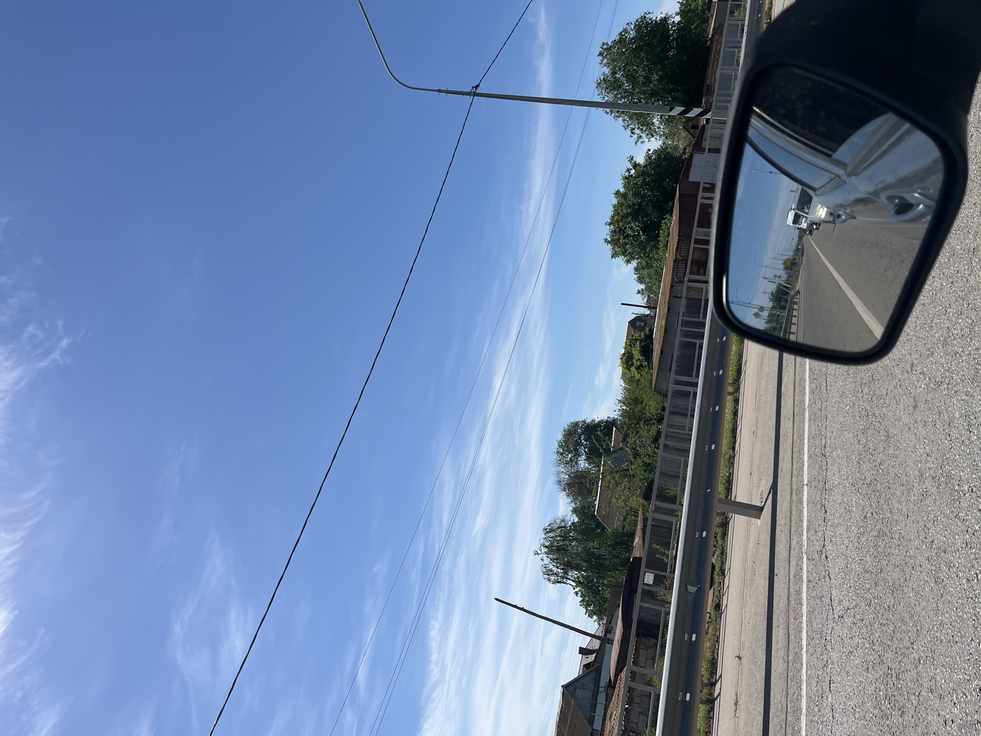 This is a photograph taken from a moving vehicle, likely a car, on a sunny day. The perspective is from the driver's seat, looking forward and slightly to the right, with a side mirror in the foreground reflecting traffic behind. The sky is a bright blue with wispy white clouds. Power lines are visible crisscrossing the sky.
In the mid-ground, there is a road with a guardrail separating it from the embankment on the left. Beyond the guardrail, there are buildings with corrugated metal roofs, some trees, and a wooden fence. A tall, thin utility pole is also visible. The reflection in the side mirror shows a line of vehicles on the road, including a white van and other cars, traveling in the same direction as the photographer. The road surface appears to be asphalt, with visible cracks and white lane markings. The environment suggests a semi-rural or suburban setting. The location is identified as Bakhtemir, Russia.