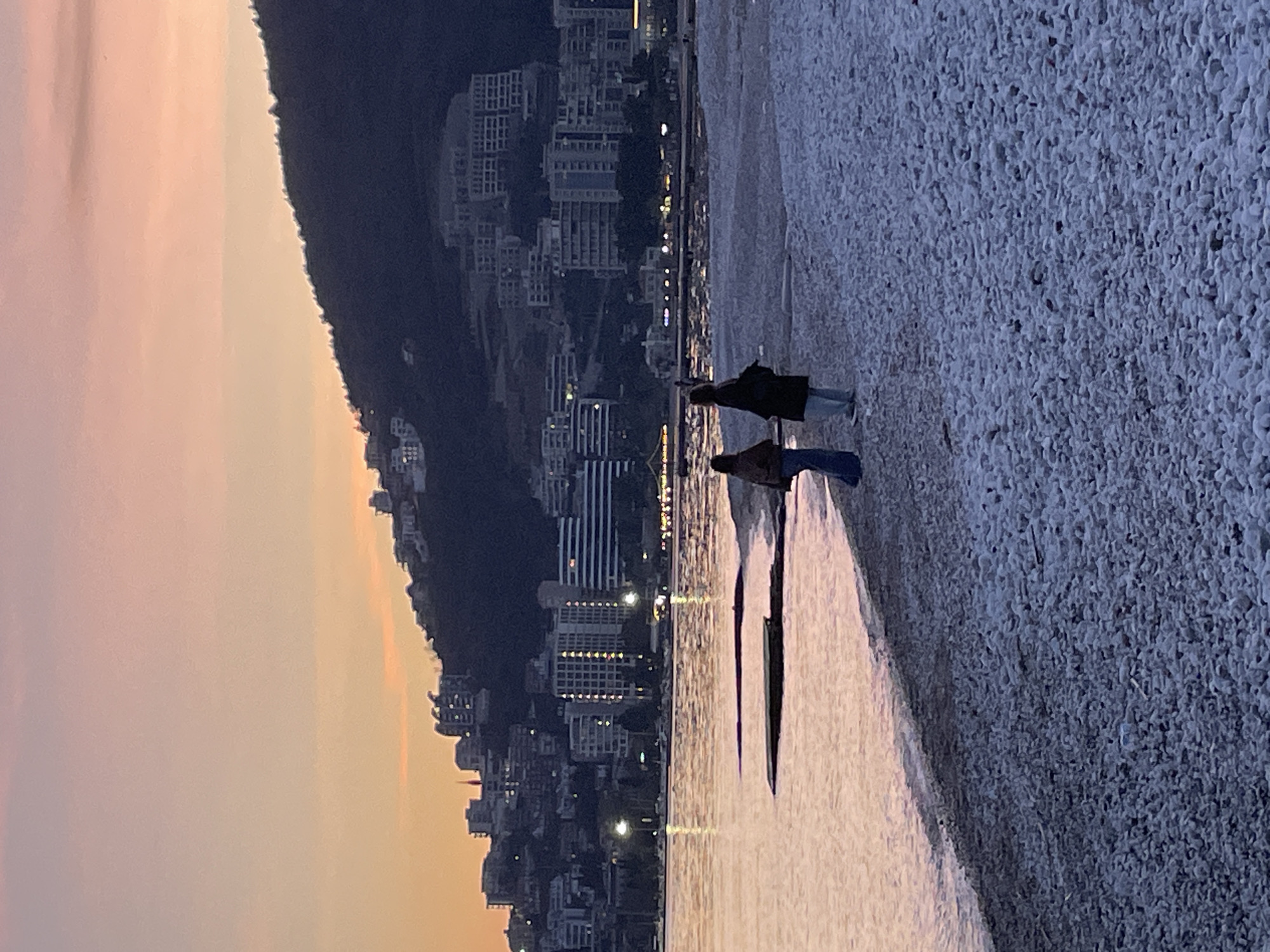 The image depicts a coastal scene in Budva, Montenegro, at dusk. A pebble beach occupies the foreground, extending from the bottom of the frame to approximately the mid-ground. The pebbles are predominantly light grey and white. To the left, the calm water of the sea meets the beach, reflecting the twilight sky's soft orange, pink, and purple hues. Several dark, low-lying structures, possibly remnants of jetties or submerged rocks, are visible in the shallow water near the shore.
Two individuals are present on the pebble beach, positioned near the water's edge with their backs mostly to the viewer. The person on the left wears dark trousers and a light-colored top or jacket. The person on the right, appearing slightly taller, wears light trousers and a long, dark coat. Both are standing still, facing away from the camera and towards the sea and the distant city.
In the background, across the water, a prominent dark hill rises. This hillside is densely covered with numerous multi-story buildings, varying in size and architectural style, which ascend the slope. Many of these buildings and structures along the waterfront are illuminated, casting bright points of light across the cityscape and reflecting faintly on the water's surface. The sky above transitions from a warm orange-yellow near the horizon to a cooler pink-lavender higher up.