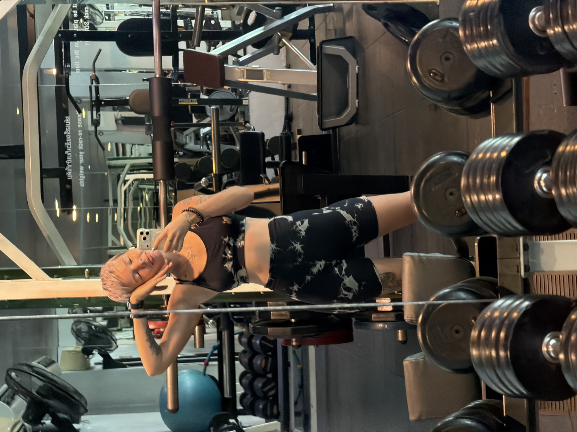 The image, rotated 90 degrees clockwise, depicts a person taking a selfie in a gym mirror, set in Thep Krasatti, Thailand. The subject is a woman with short, light-colored hair, wearing a black and white tie-dye sports bra and matching biker shorts. She has several tattoos on her left arm and chest, a nose piercing, and a necklace. She holds a smartphone up to capture her reflection, smiling serenely with her head resting on her right arm.
The background, seen through the mirror's reflection, reveals a typical gym environment. Rows of heavy dumbbells are stacked on a rack to the right, some of which are partially visible in the foreground. Various pieces of workout equipment, including a leg press machine and other weight training apparatus, are visible further back. A blue exercise ball and a black fan are on the floor to the left. The artificial lighting suggests an indoor setting, possibly during the evening or night.
Visible text in the reflection includes Thai characters, such as "นลิเกย์เบกเริ่มแรกเมร", which appears to be part of a sign. Other discernible text elements are "LSCK 201", "siq ed ta", and "6926 LG-LOCK λoπι Μειθμια", suggesting labels or other signage. The brand "POWERTEC" is clearly visible on a piece of equipment in the background.
