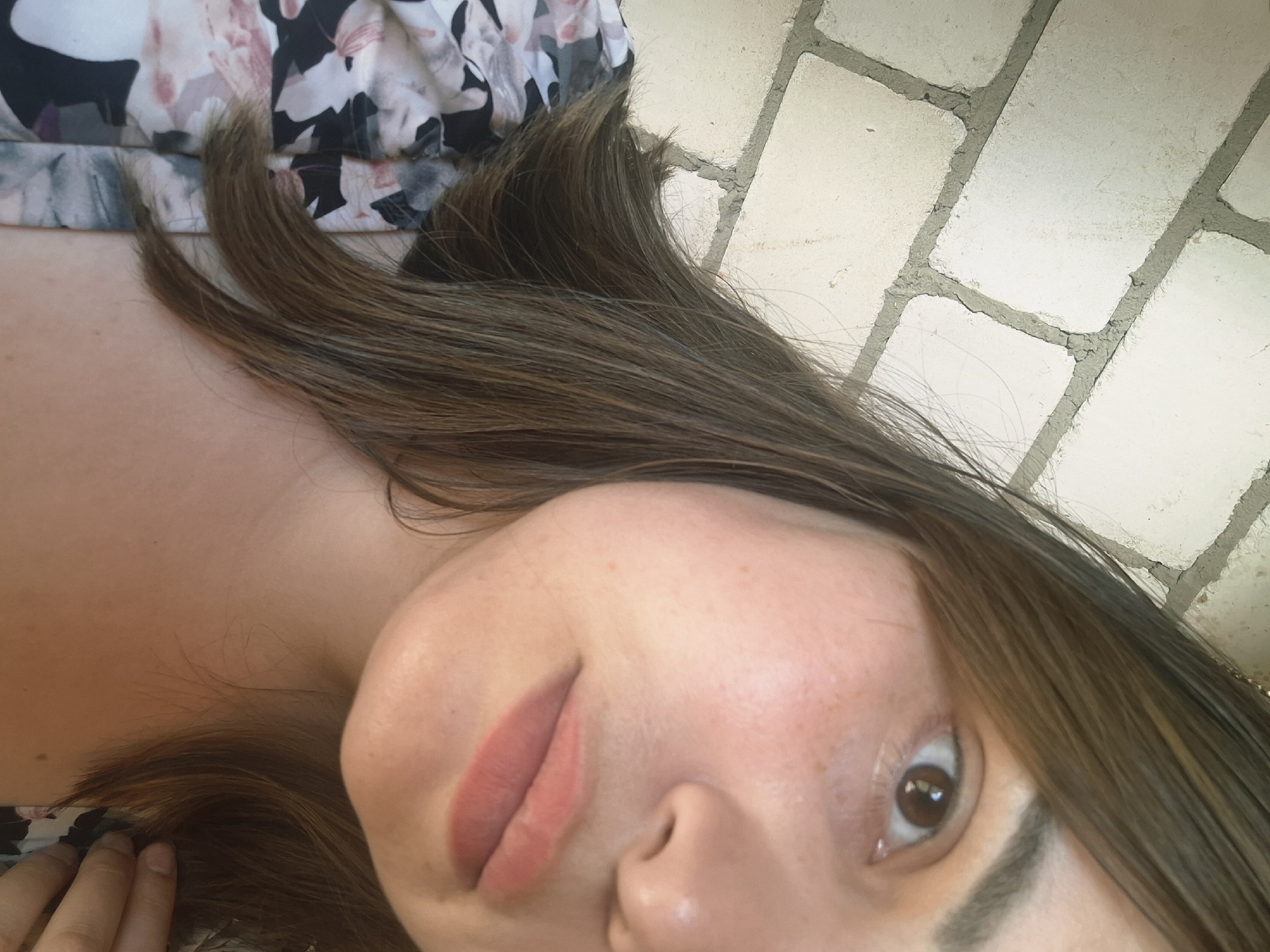 A close-up, slightly angled portrait shows a person with fair skin, brown hair, and brown eyes. The subject is lying down, with a floral-patterned garment visible near their shoulder and hand. The background features a light-colored brick or tile surface with gray grout lines. The image does not provide enough information to identify a specific location in Амзя, Russia, or to determine any actions or interactions occurring.