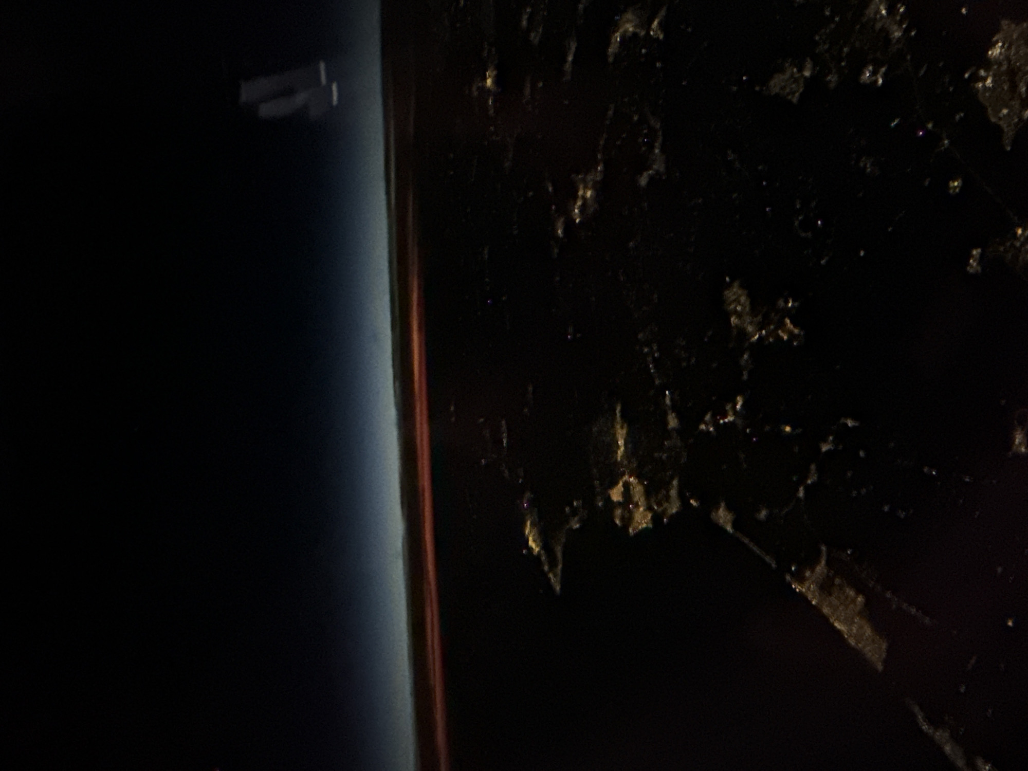 The image appears to be a nighttime shot from space, possibly taken from the International Space Station. The Earth's curvature is visible, with a blue atmospheric haze along the left side. A faint object, possibly a satellite or part of the space station, is visible in the upper left. On the right side, the dark surface of the Earth is visible, with sparse points of light suggesting city lights. The location might be Sofia, Bulgaria, based on the distribution of lights in the dark area and its geographic coordinates.