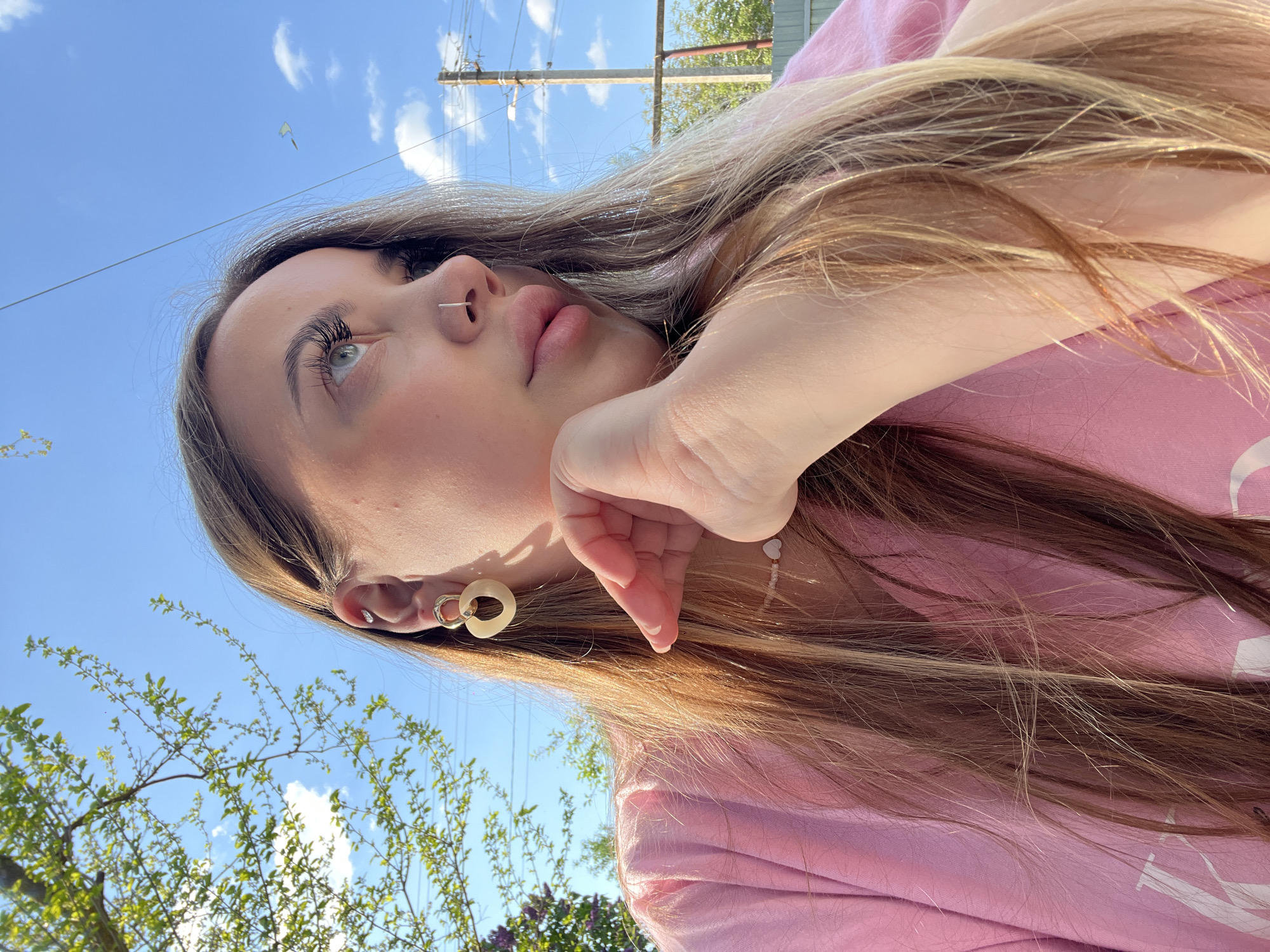 A young woman lies with her head angled upwards, facing left. She has long blonde hair with darker roots. She wears a pink shirt. Her left hand is held up near her face. A silver nose ring is visible in her left nostril. She wears a light-colored earring in her left ear. Behind her are trees and a bright blue sky with some clouds. Overhead electrical lines and poles are also visible. No specific details indicate the location as Novopokrovskaya, Russia.