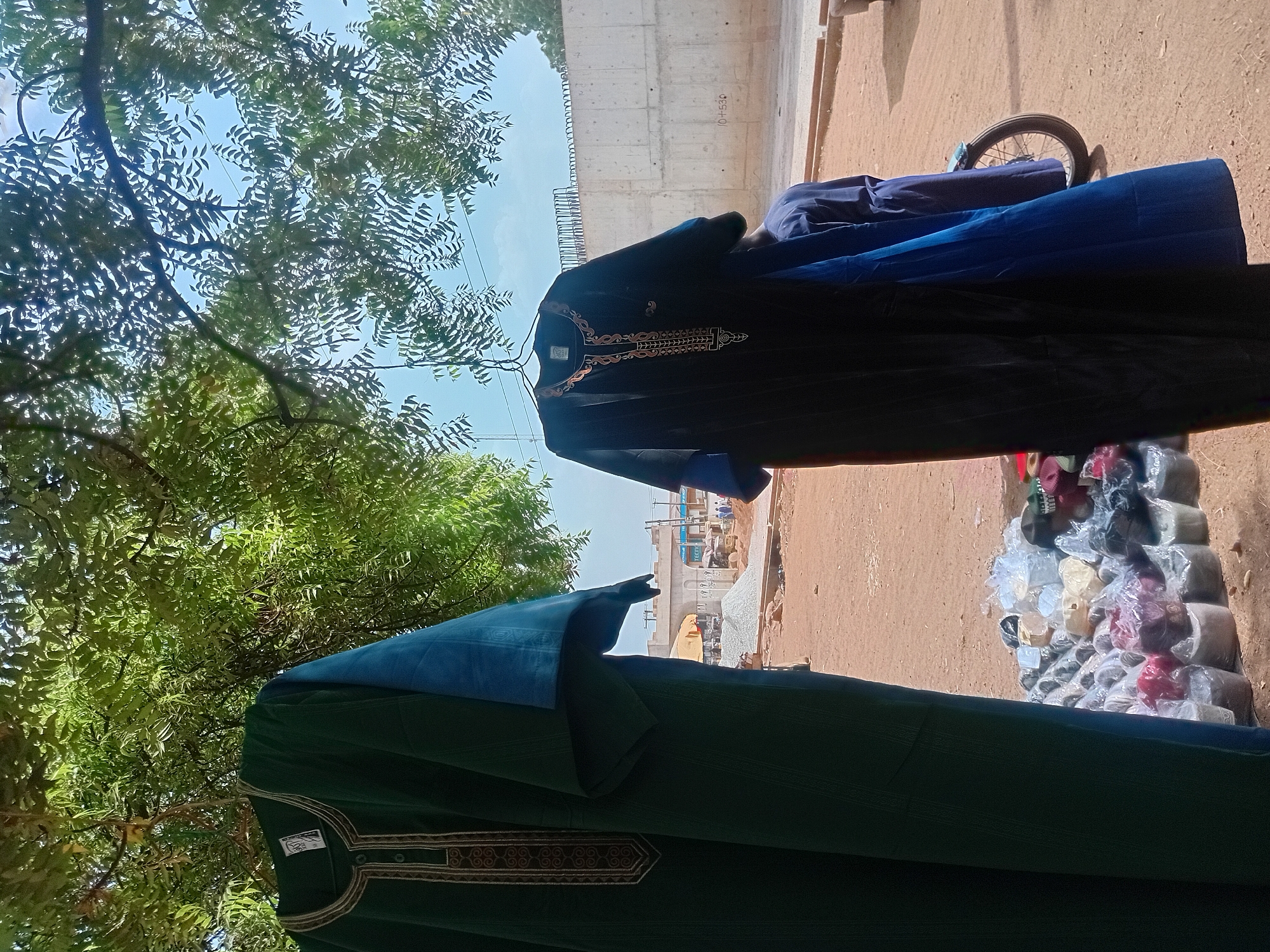 An outdoor market or street vending scene is captured under a bright, sunny sky in what appears to be Minna, Nigeria. The image is taken during the day, with clear blue skies visible through the dense canopy of green trees that dominate the upper left portion of the frame, suggesting shade.
In the foreground, two traditional long-sleeved garments are displayed vertically, likely hung for sale. The garment on the left is dark green with intricate brown and beige embroidery around the neckline and placket, featuring a small white tag with illegible text. The garment on the right is black, adorned with detailed gold and brown embroidery. Below the black garment, a roll of dark blue fabric is visible, with a lighter blue fabric draped over it.
The ground beneath the displayed items is dirt-colored. On the right side, a pile of smaller merchandise is stacked on the ground, consisting of numerous items, many individually wrapped in clear plastic. These appear to be various types of headwear, such as kufis or small hats, in assorted colors including white, beige, red, and black. Part of a bicycle wheel is partially visible behind these items.
In the background, a large concrete structure, possibly an elevated road or bridge under construction, spans horizontally. Faint markings like "Ø +530" are visible on its surface. Further in the distance, behind the construction, other buildings, including one with an archway, can be seen, indicating an urban or semi-urban setting. The scene depicts a typical commercial activity in a bustling environment.