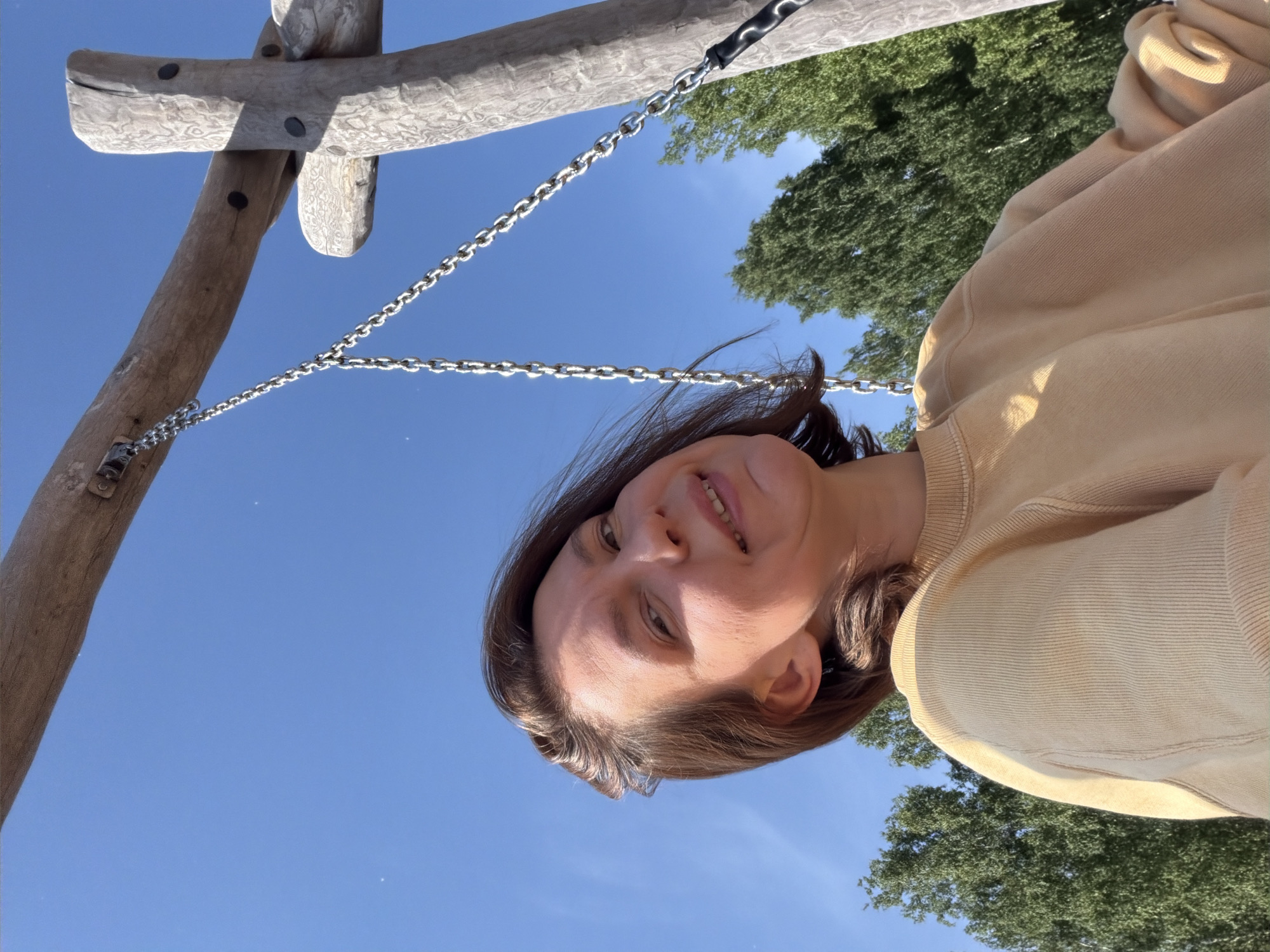 A woman with brown hair and fair skin is captured in a low-angle, selfie-style shot from a swing. She is wearing a light yellow crewneck sweatshirt. The background reveals a clear blue sky and lush green trees, suggesting an outdoor setting. A wooden swing set frame with metal chains is visible in the foreground. The image appears to have been taken in Chaikovsky, Russia, likely during daylight hours given the bright illumination.