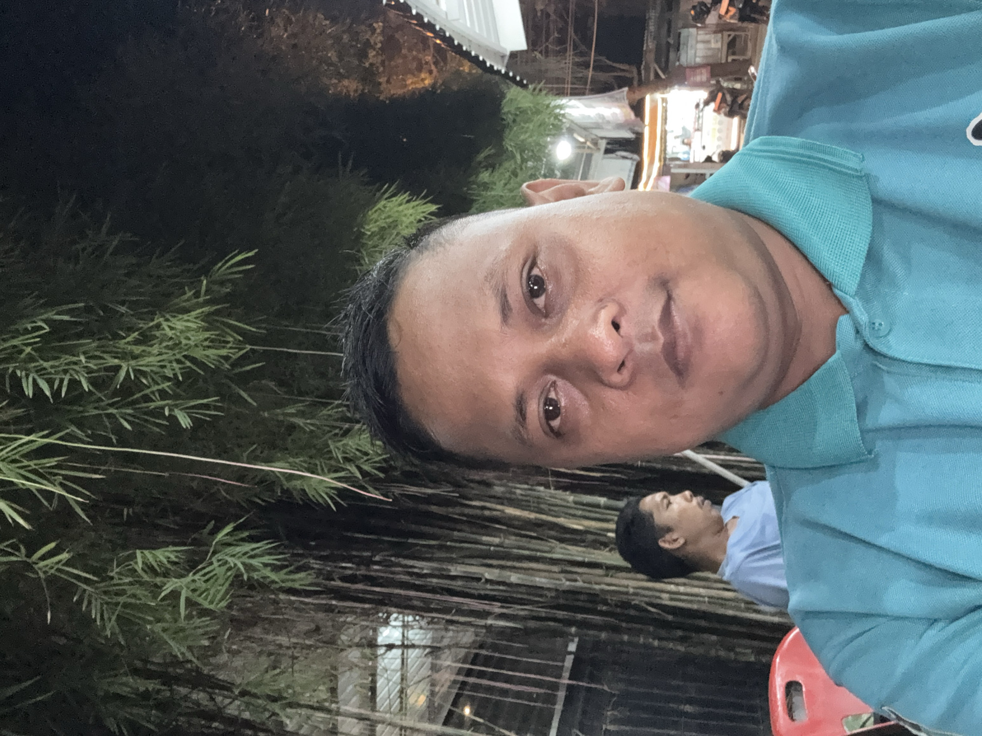 The image shows a man in a blue shirt taking a selfie. Another person, also wearing a blue shirt, is visible in the background, sitting in a red chair. The background includes what appears to be bamboo stalks and some nighttime urban lights. The location is potentially Lhokseumawe, Indonesia.