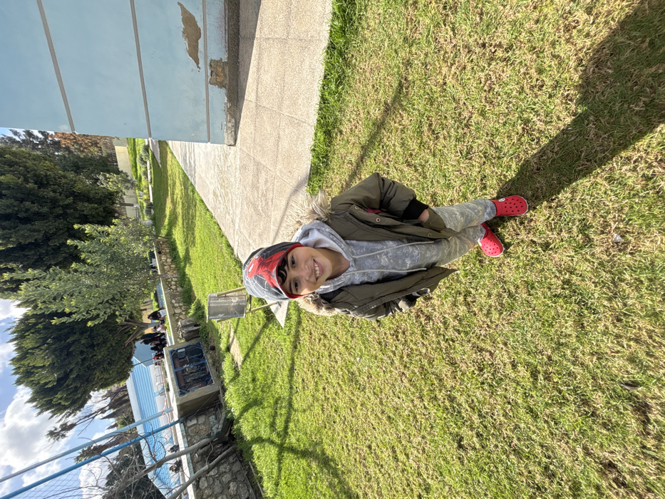 A young male subject stands centrally on a sunlit grassy lawn in Meknes, Morocco. The subject, a child, faces the camera directly, displaying a smile. He is dressed in a grey, red, and black beanie with a black symbol, a grey hooded sweatshirt, an olive green winter jacket featuring a fur-trimmed hood, grey patterned sweatpants, and red Crocs. His hands are placed inside his jacket pockets. A prominent shadow from the subject is cast across the grass behind him to the left. To the subject's right, a concrete pathway runs parallel to a light blue building, the lower section of which exhibits significant paint peeling and exposed material. To the left, a stone retaining wall separates the foreground lawn from a lower elevation. Beyond this wall, a blue and white structure, consistent with a sports court boundary or net support, is visible. Further in the background, a group of approximately five to seven individuals, largely obscured, are present on a grassy slope beneath several tall evergreen trees. A silver-colored metal trash receptacle stands on the lawn between the subject and the retaining wall. The sky overhead is partially cloudy.