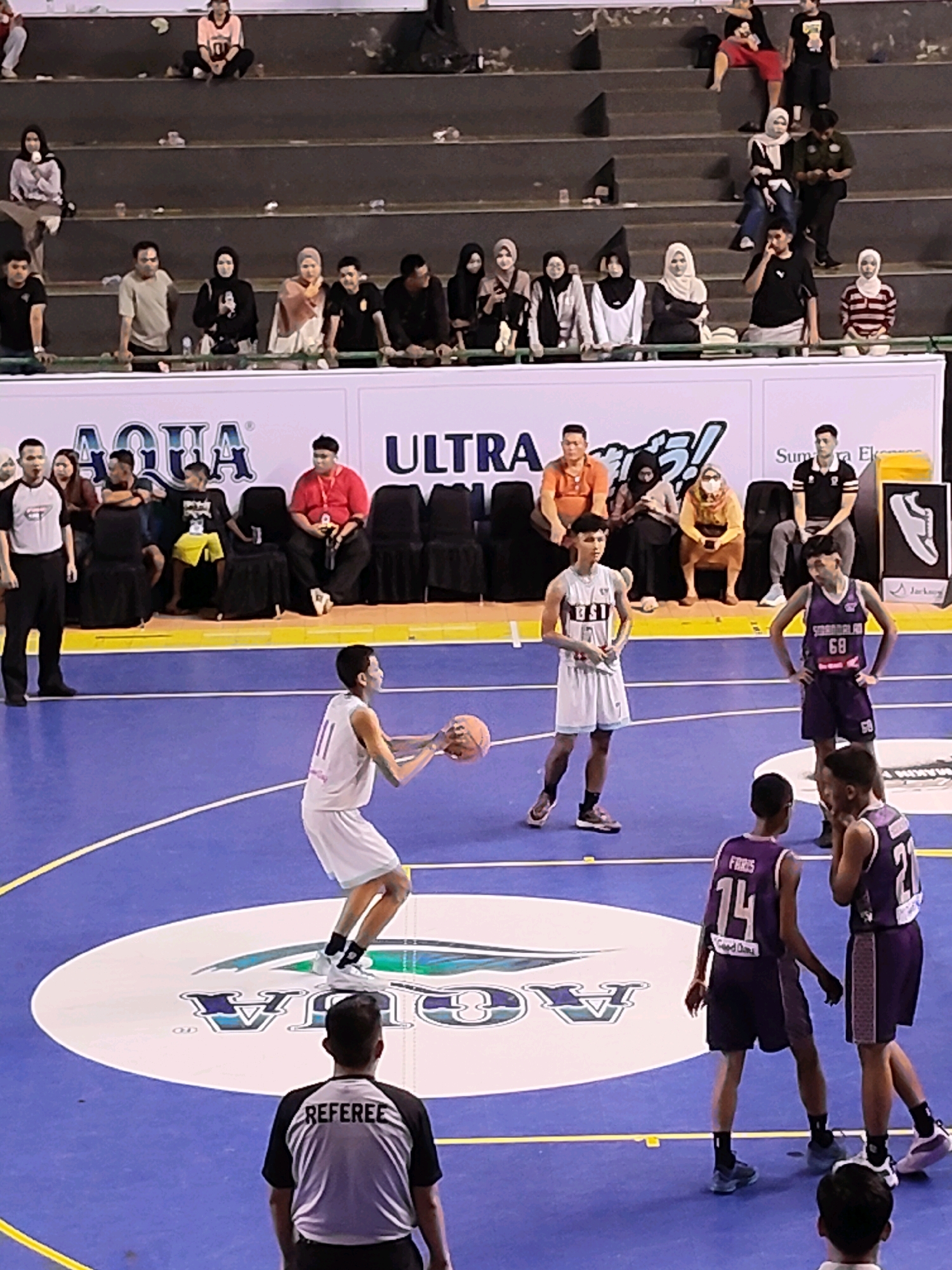 The image captures a basketball game in progress, likely indoors given the presence of bleacher seating and a banner advertising "AQUA" and "ULTRA." The court is a vibrant blue with white and yellow lines, and a circular logo for "AQUA" is visible at center court.
On the court, several young men in basketball uniforms are actively playing. One player in a white uniform, with the number 11, is in the process of shooting a basketball. Another player in a purple uniform, wearing number 68, stands with his hands on his hips, observing. Two other players in purple uniforms are also on the court, one with "FARIS" and the number 14 visible on his jersey, and another with the number 21. A referee, dressed in a black and white striped shirt with "REFEREE" printed on the back, stands on the court, watching the game.
In the background, spectators are seated in tiered bleachers. Some are wearing headscarves, suggesting a cultural context. The overall atmosphere appears to be that of a competitive sporting event.
While the image itself does not contain explicit identifiers for the location in Palembang, Indonesia, the presence of the "AQUA" and "ULTRA" branding, commonly seen in Indonesian advertisements, along with the visible cultural attire of some spectators, strongly suggests an Indonesian setting. Given the context of a basketball game with this branding, it's plausible this event is taking place in a sports facility or stadium within Palembang, Indonesia.