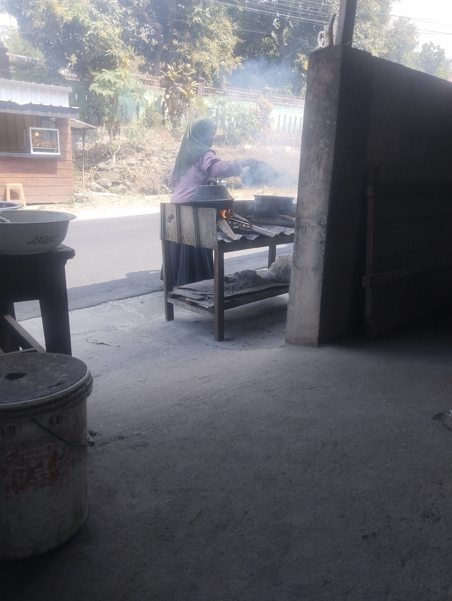 A woman wearing a green headscarf and a pink shirt stands in front of a makeshift stove with a pot over an open fire. Smoke rises from the fire, partially obscuring her and the surroundings. She appears to be tending to the cooking. To her left, a wooden structure with a white roof and a window displays some items inside. The scene is set outdoors, with a paved road in the background and vegetation. The ground in the foreground is dusty or sandy. To the far left, a large, grey, weathered bucket is visible. The image has a grainy quality and is overexposed in some areas. The location is identified as Kembang Kerang, Indonesia.