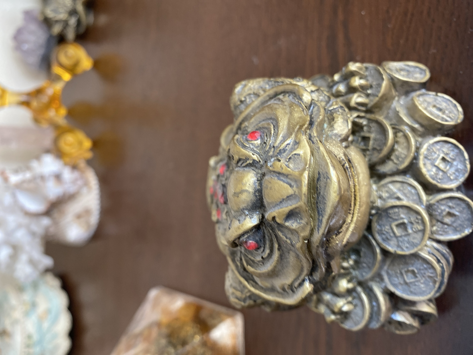 A close-up, eye-level shot shows a golden, frog-shaped ornament on a brown wooden surface. The frog has red eyes and is adorned with red markings on its head. Its base is decorated with numerous small, circular coins, also in a golden hue. In the background, out of focus, are various decorative items, including what appear to be crystals and shells. The lighting is soft, casting subtle shadows. There is no indication of the city of Brest, Belarus, in this image.
