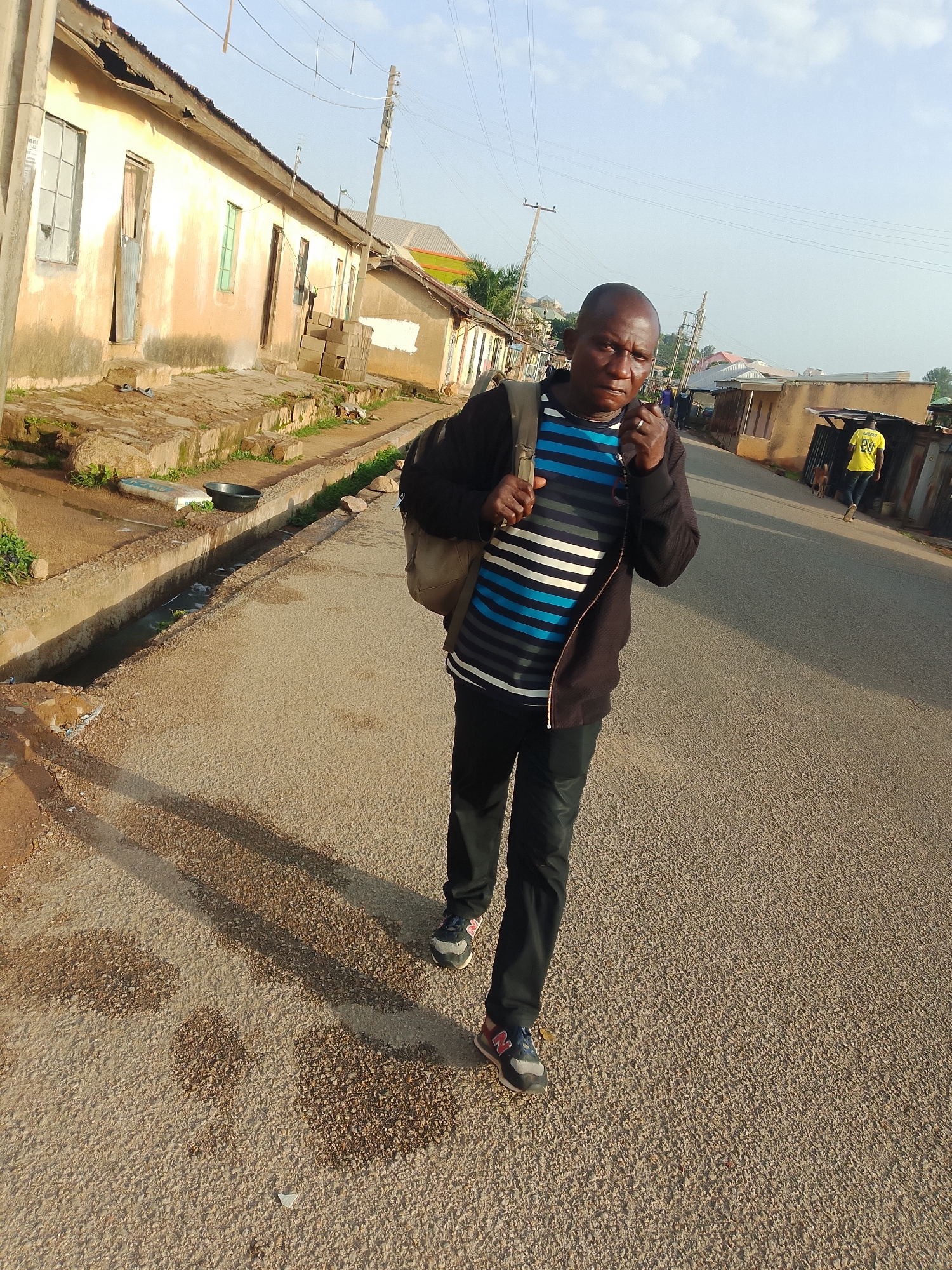 The image depicts a street scene in Jos, Nigeria. A man in his 40s with a shaved head and medium complexion is walking towards the camera, wearing a black zip-up jacket over a horizontally striped blue and white t-shirt, black pants, and multi-colored athletic shoes. He is carrying a beige backpack. To his left, a drainage channel runs alongside the road, containing some water and debris. Buildings with worn facades, some with corrugated iron roofs, line the left side of the street. A second man, wearing a bright yellow t-shirt, is walking away from the camera in the distance on the right side of the road, accompanied by a dog. The road surface is asphalt with visible gravel. The sky is partly cloudy.