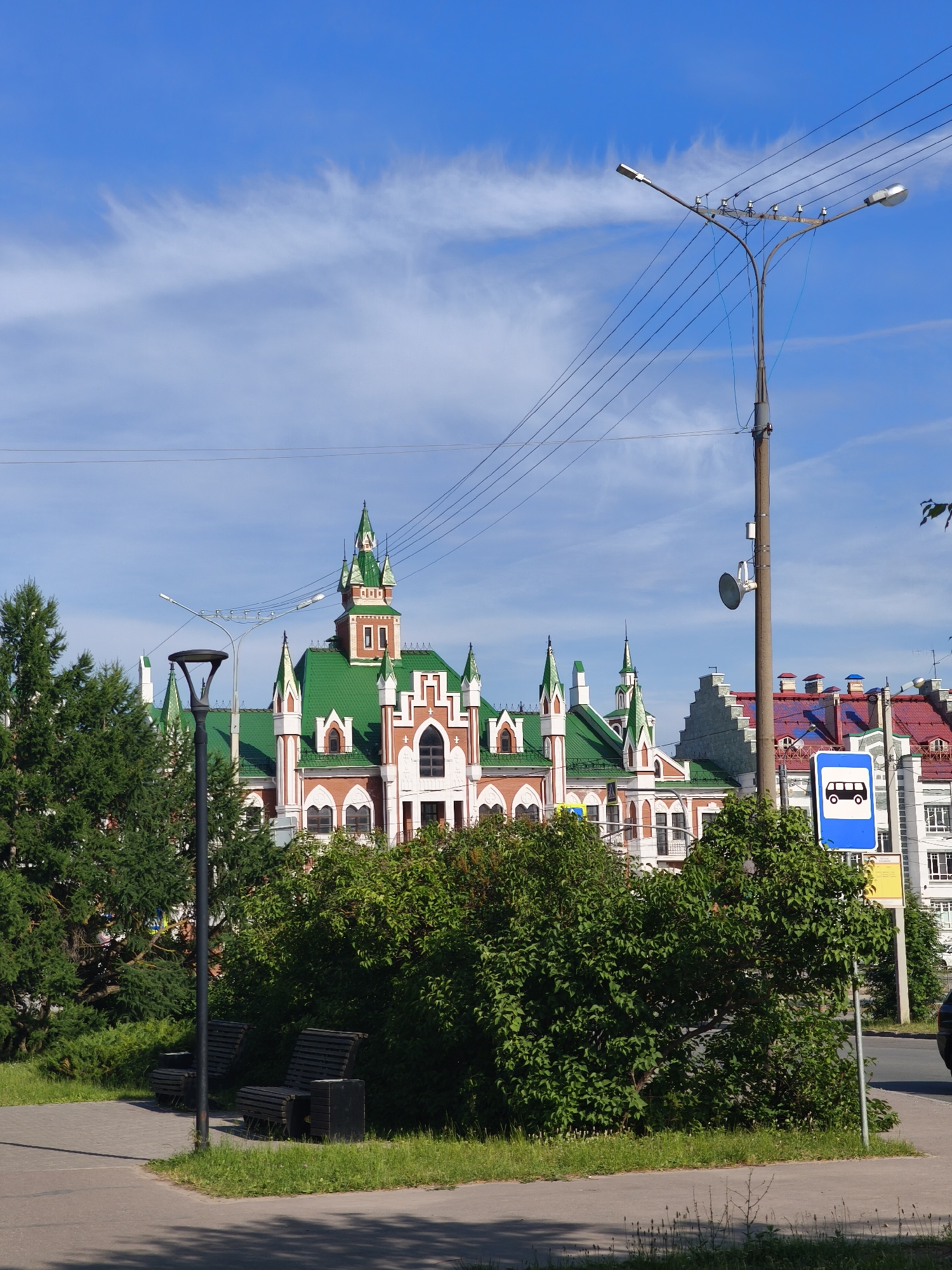 The image captures a sunny day in Yoshkar-Ola, Russia, with a blue sky dotted with wispy clouds. In the foreground, a paved walkway borders a grassy area with two dark wooden benches. A tall, dark lamppost stands to the left of the benches, and another lamppost with multiple lights and overhead wires is visible to the right. A blue bus stop sign is prominently displayed on a pole near the right edge of the frame. Behind the greenery and lampposts, a striking building with red brick walls and a vibrant green, multi-peaked roof dominates the background. This distinctive architecture, possibly a historic train station or public building, features white decorative elements and arched windows. Further in the distance, other buildings with similarly colorful roofs can be seen. The overall scene suggests a public space in the city, likely a park or square adjacent to a transport hub.